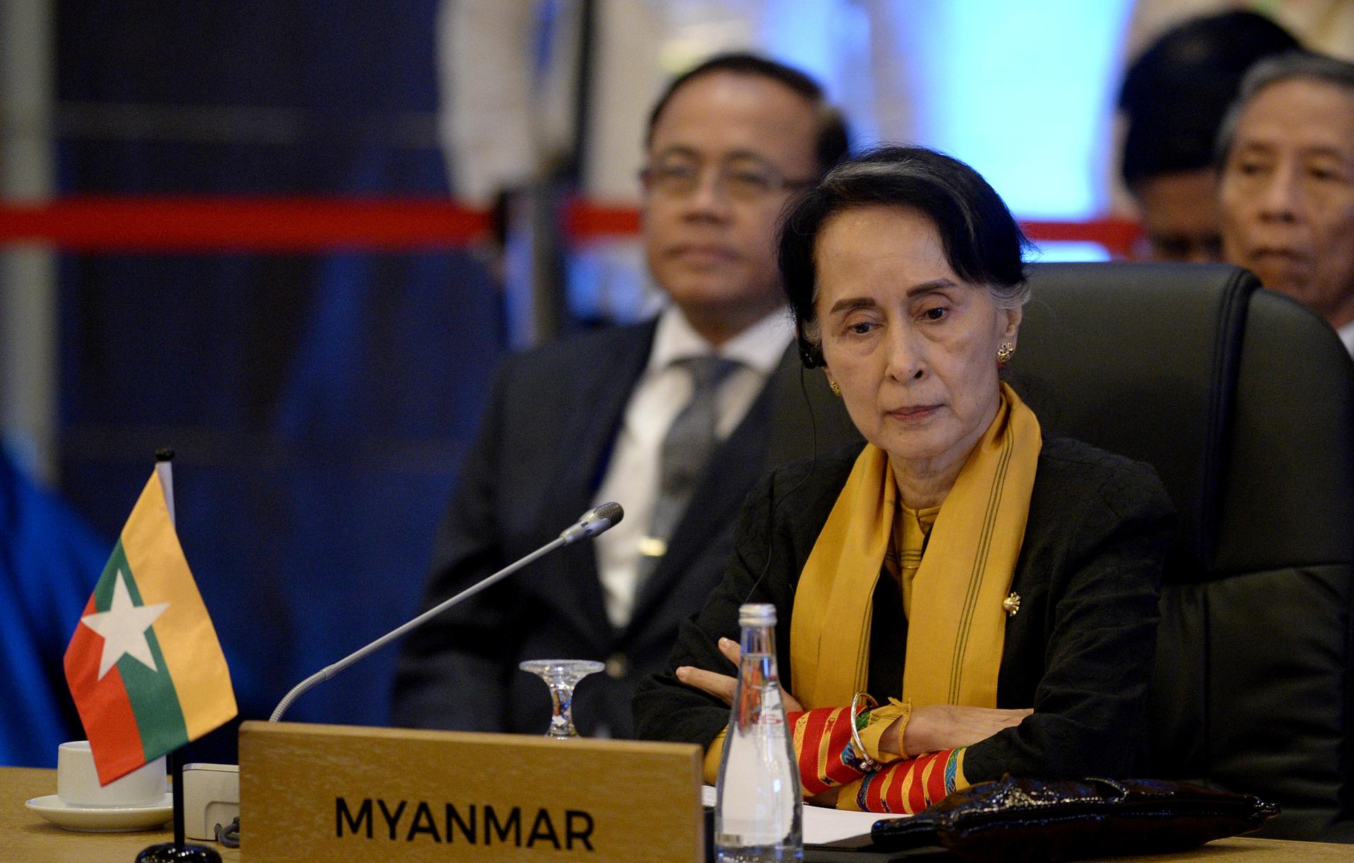 Aung San Suu Kyi looks down while sitting at a table behind a desktop placard with 