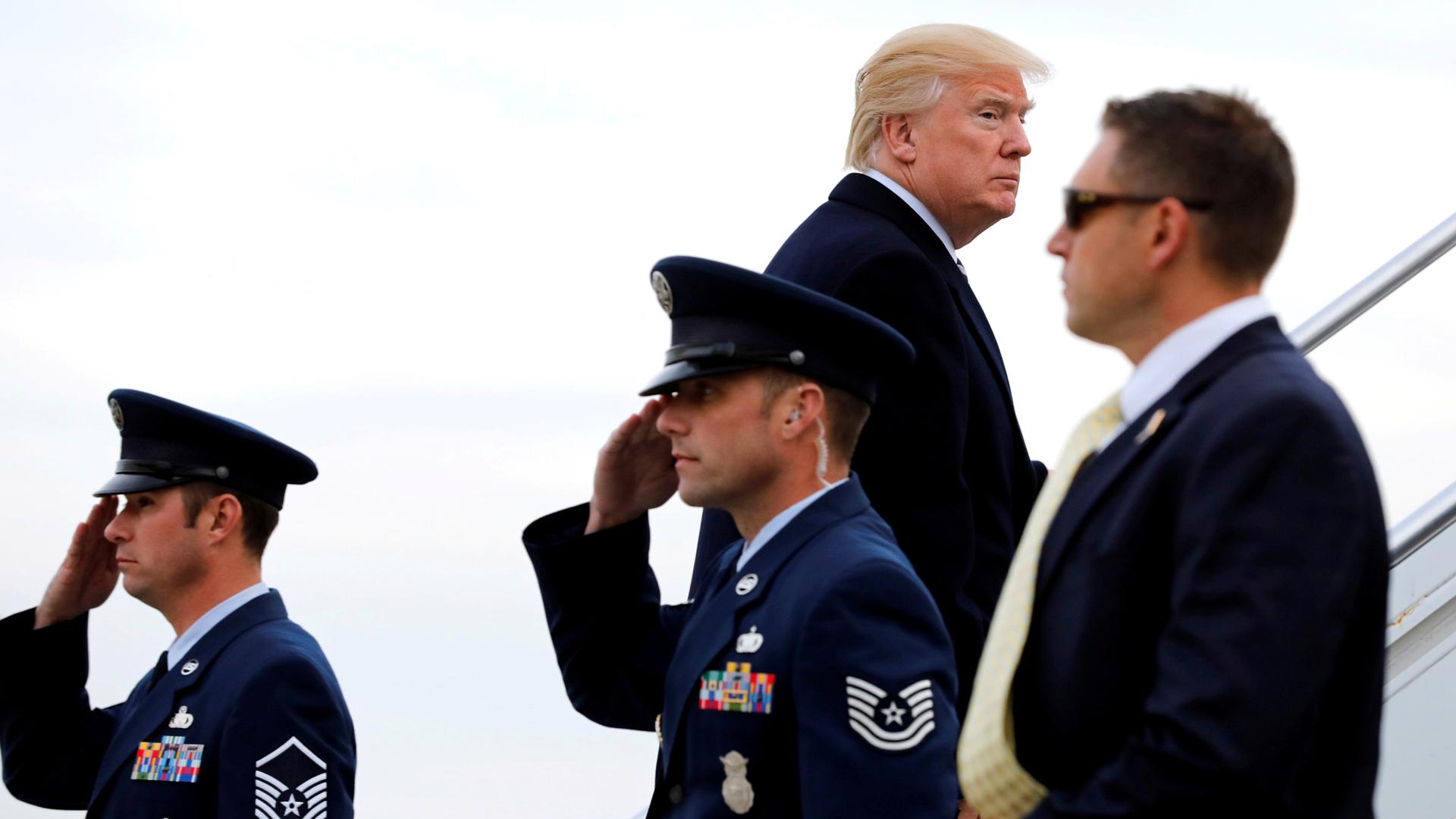President Donald Trump looks to his right while walking up the stairs to Air Force One with two Air Force officials soluting in the foreground.