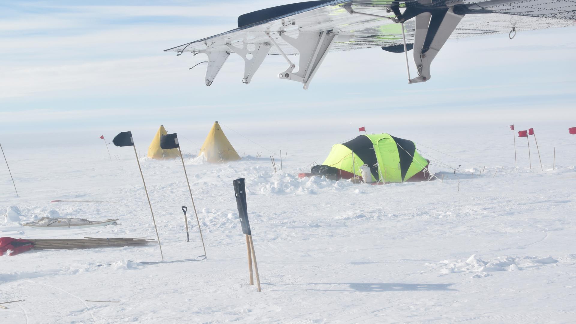 Two yellow tents and a larger dome tent are staked into the snow. At the top of the image is a wing of a tiny prop plane. 
