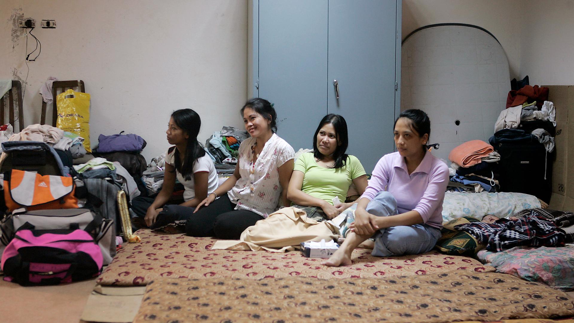 Four women sit crosslegged on the floor of a windowless room. Around them are suitcases and bedding. A baby is wrapped in a blanket nearby.