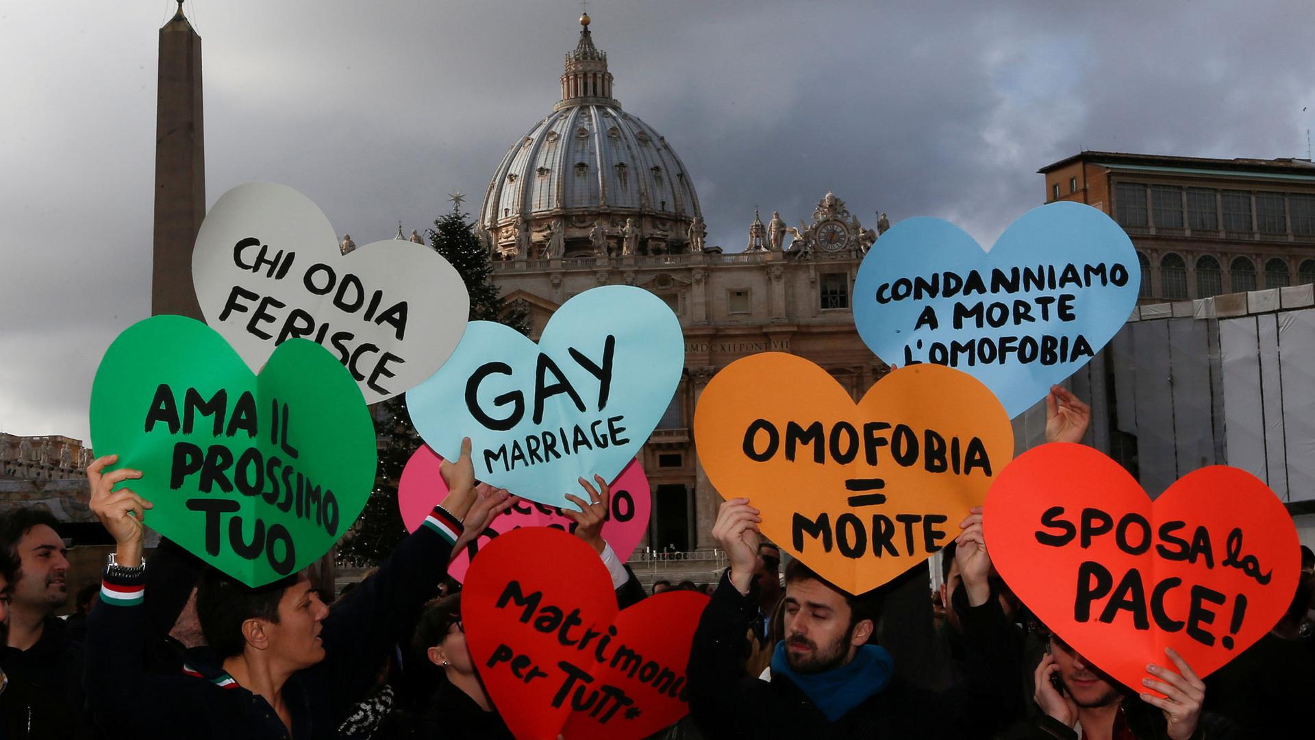 Several people are shown standing in St. Peter's square and holding heart-shaped signs.