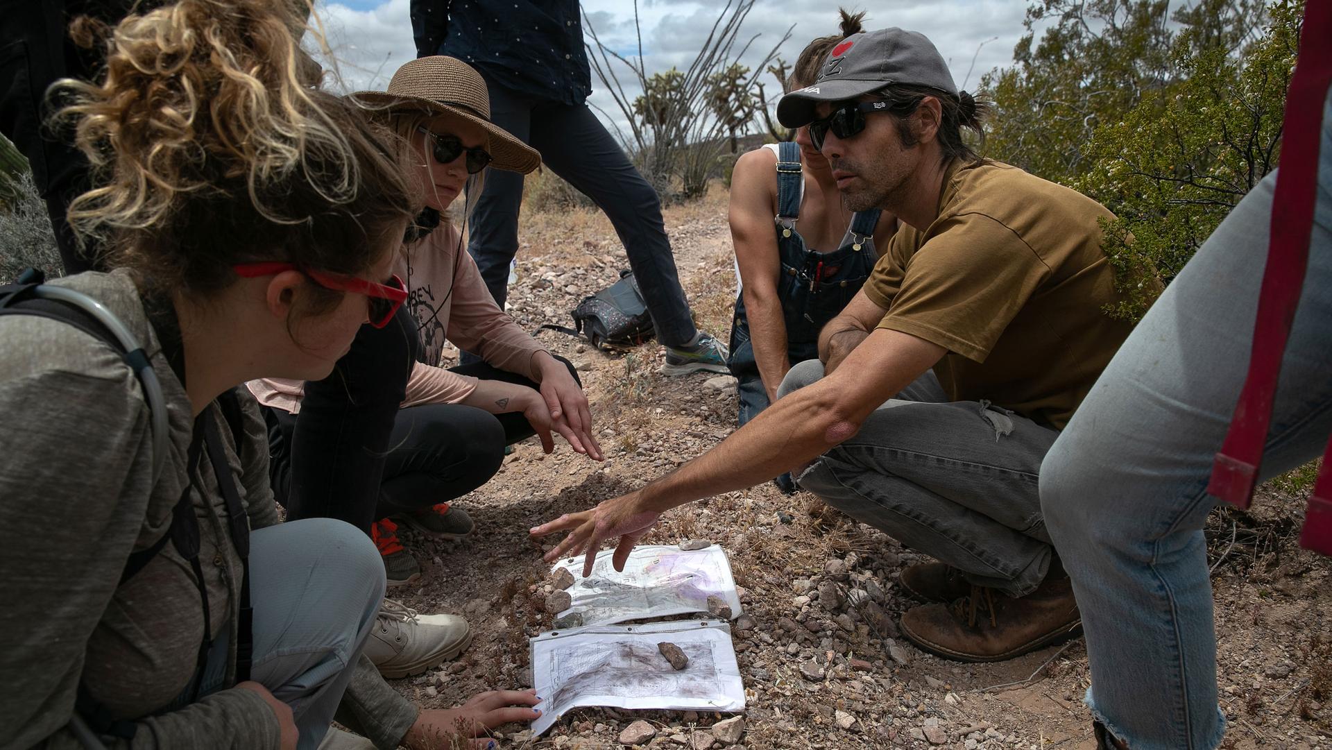 A man crouches in the dirt surrounded by others as they review a few maps. 
