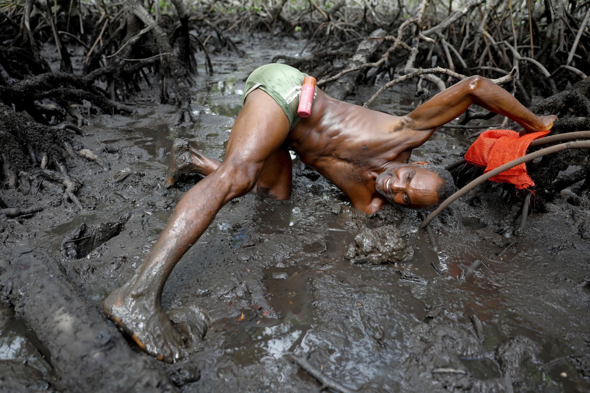A man is shown laying on mud-covered mangroves wearing only green shorts with his left arm in the mud to his shoulder.