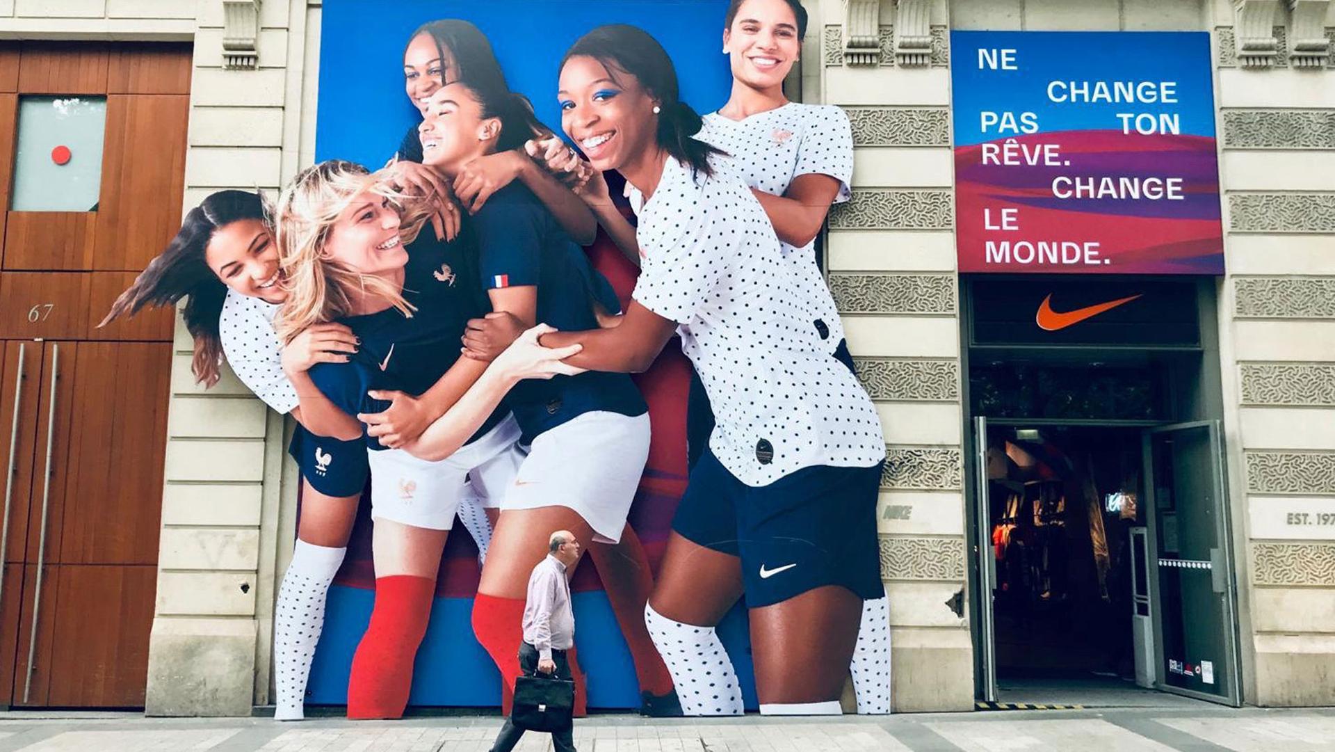 A manis shown with a briefcase walking past a billboard showing members of the French Women's World Cup soccer team hugging.