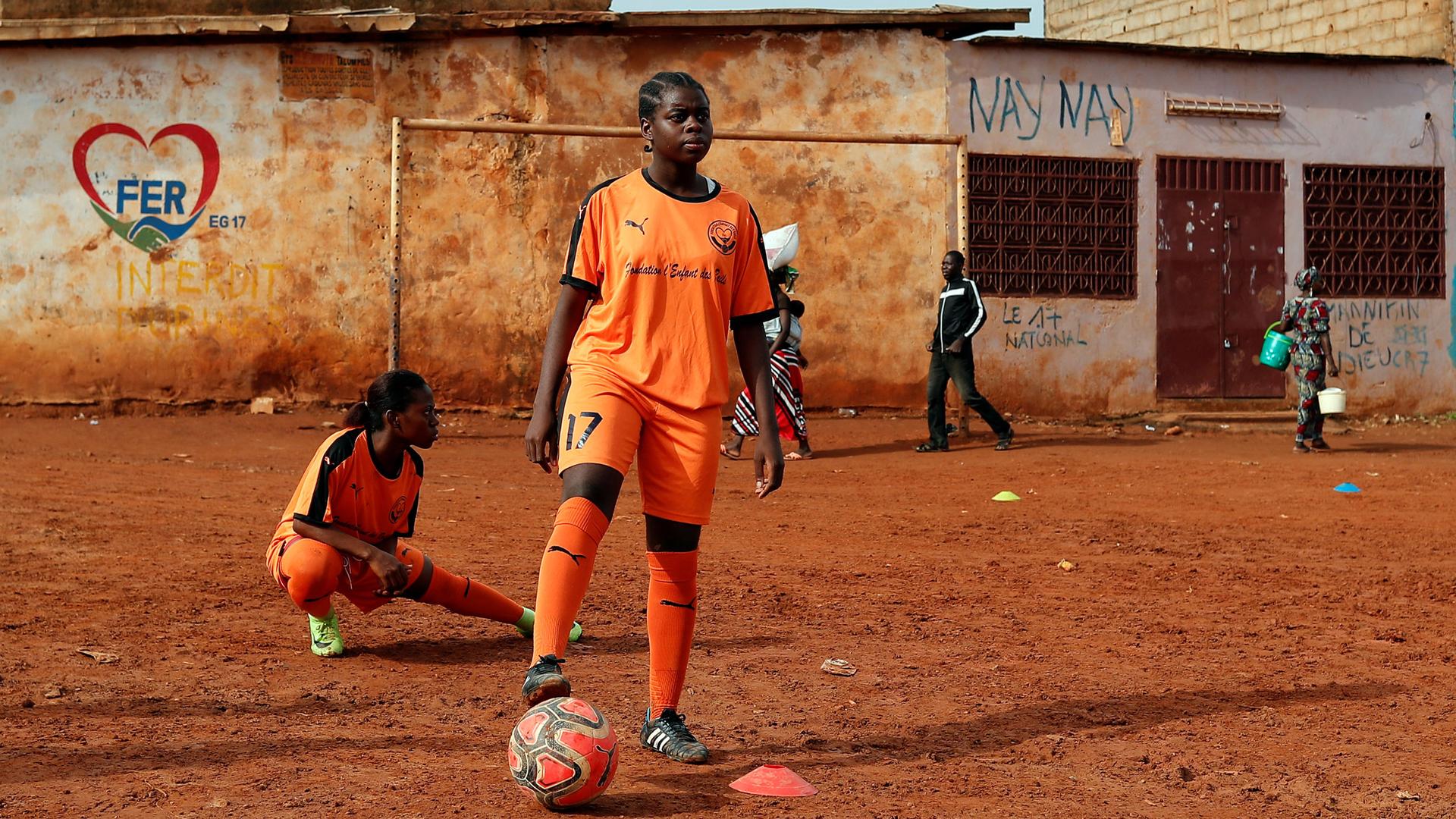 Gaelle Dule Asheri is shown standing with one foot on a soccer ball and wearing an orange uniform.