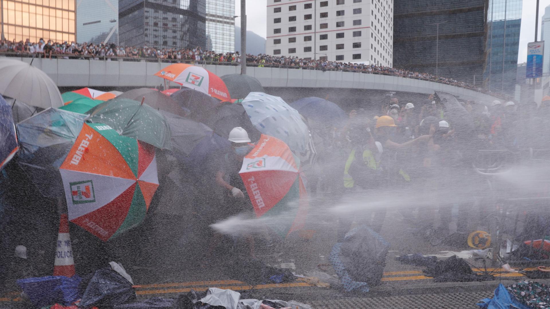 A group of protesters are shown with their umbrellas pointed down to protect themselves from water being shot across the frame of the photograph.