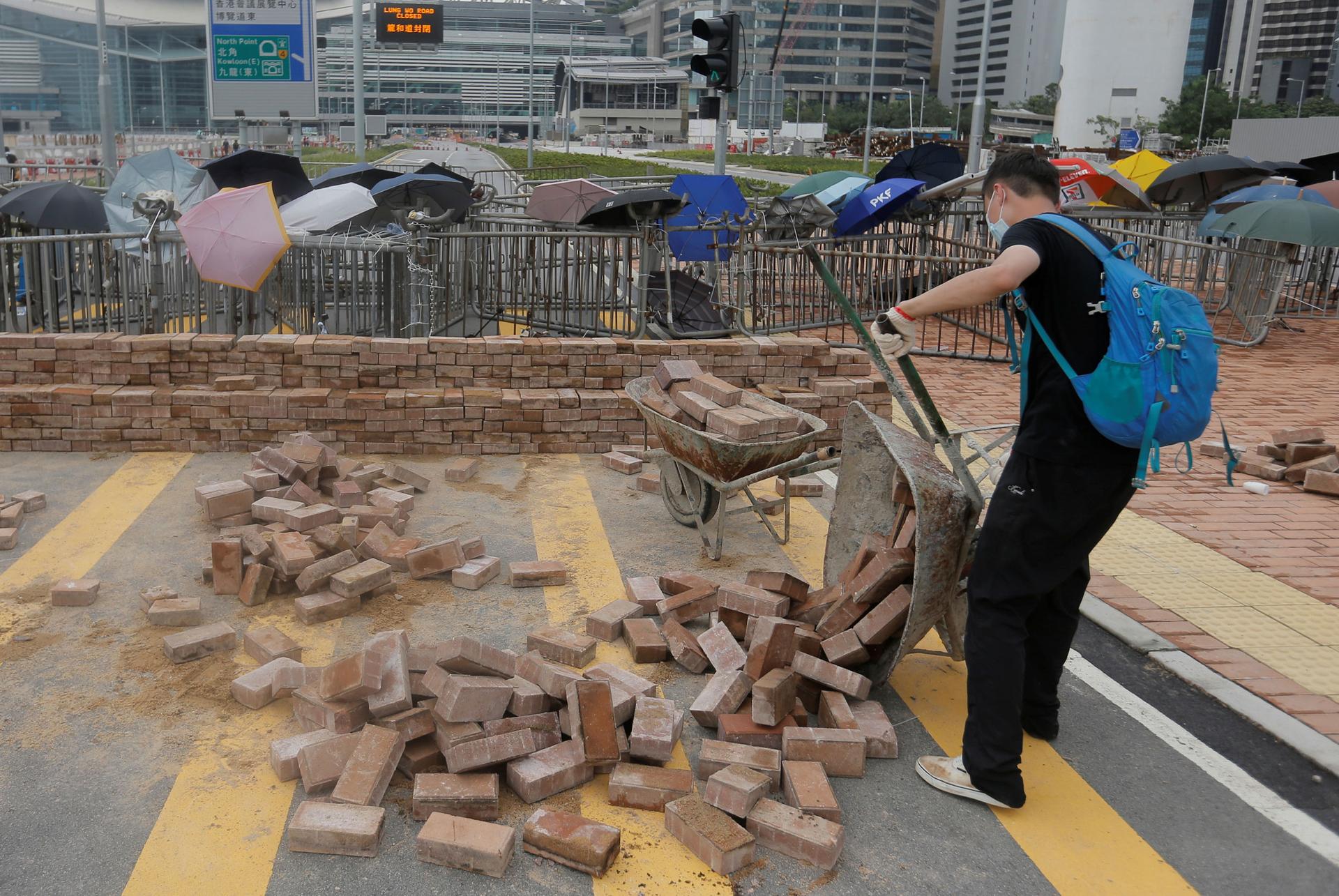 A man is shown wearing a blue backpack and dumping out a wheelbarrow of bricks onto the street.