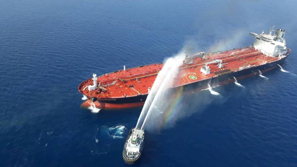 An oil tanker is shown from above with a red deck and being sprayed by water by a second, adjacent boat.