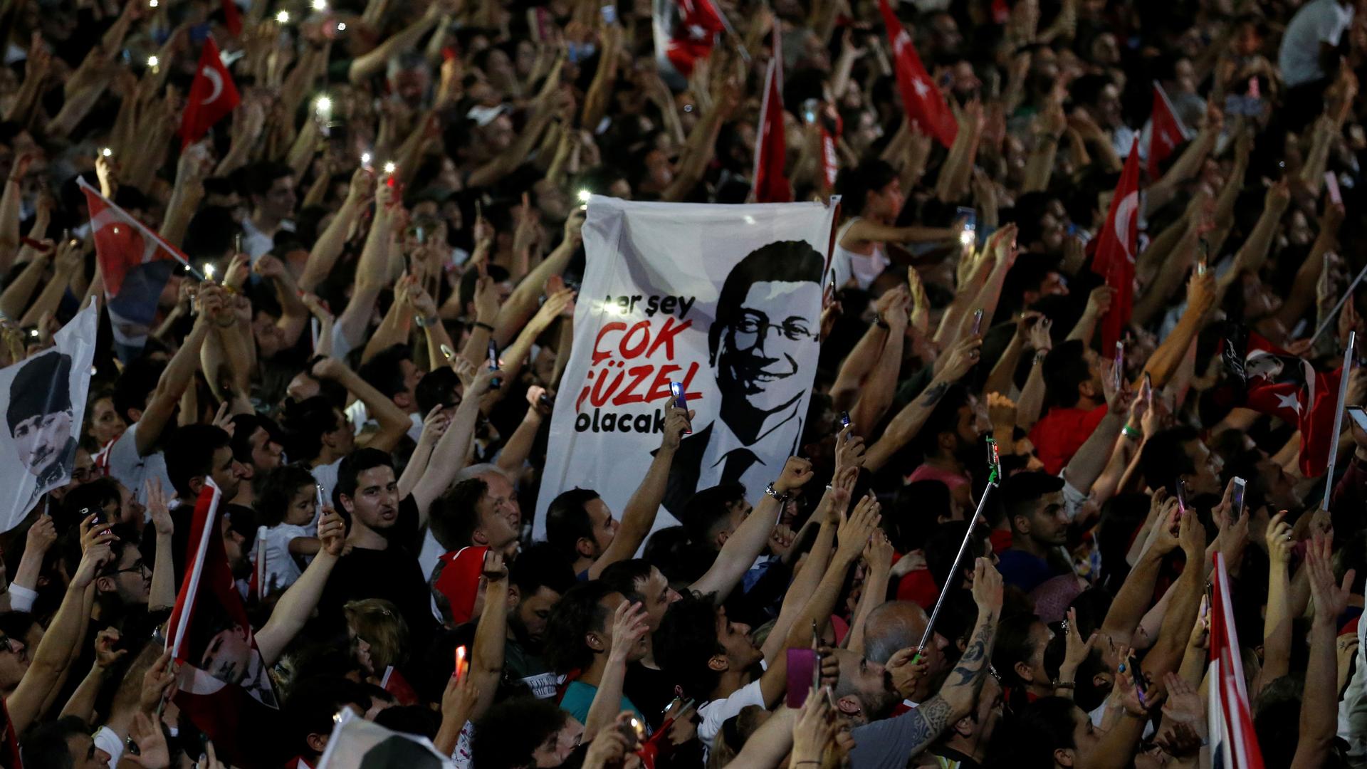 Hundres of people are shown with their hands in the air holding lighters and Turkey flags with a white sign in the middle with a portrait of Ekrem İmamoğlu on it.