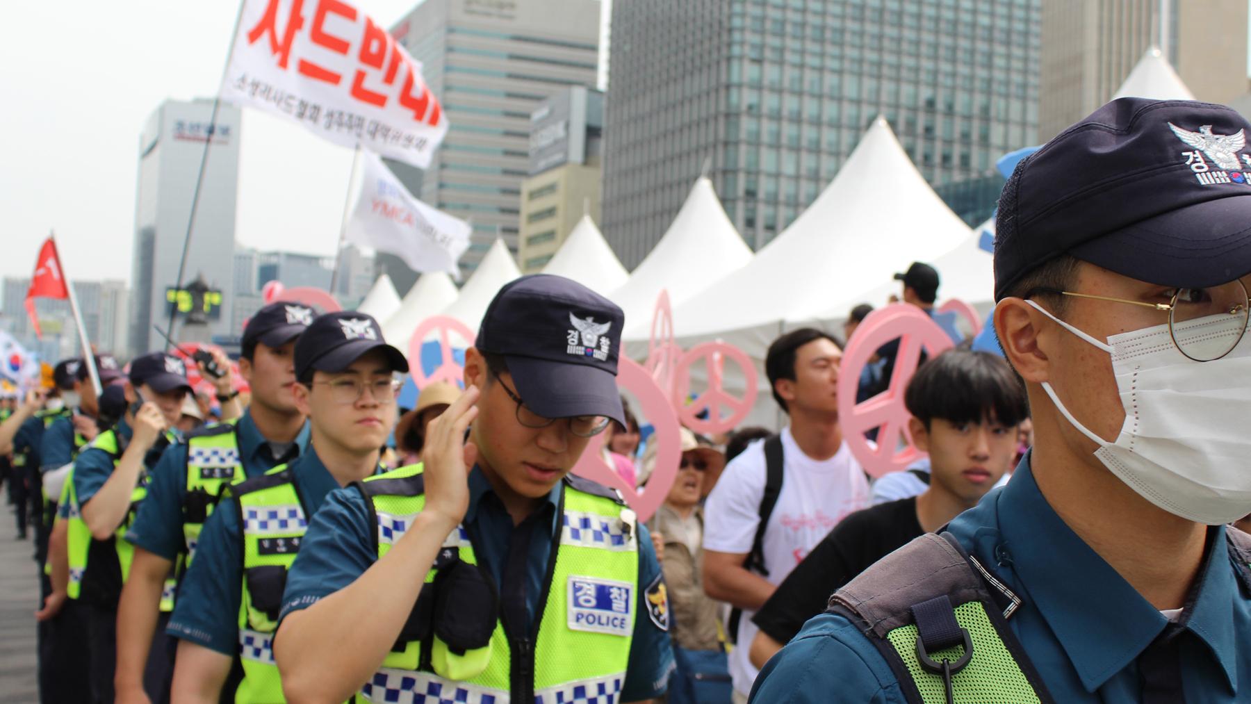Police control crowd in protest with signs with Korean letters. 