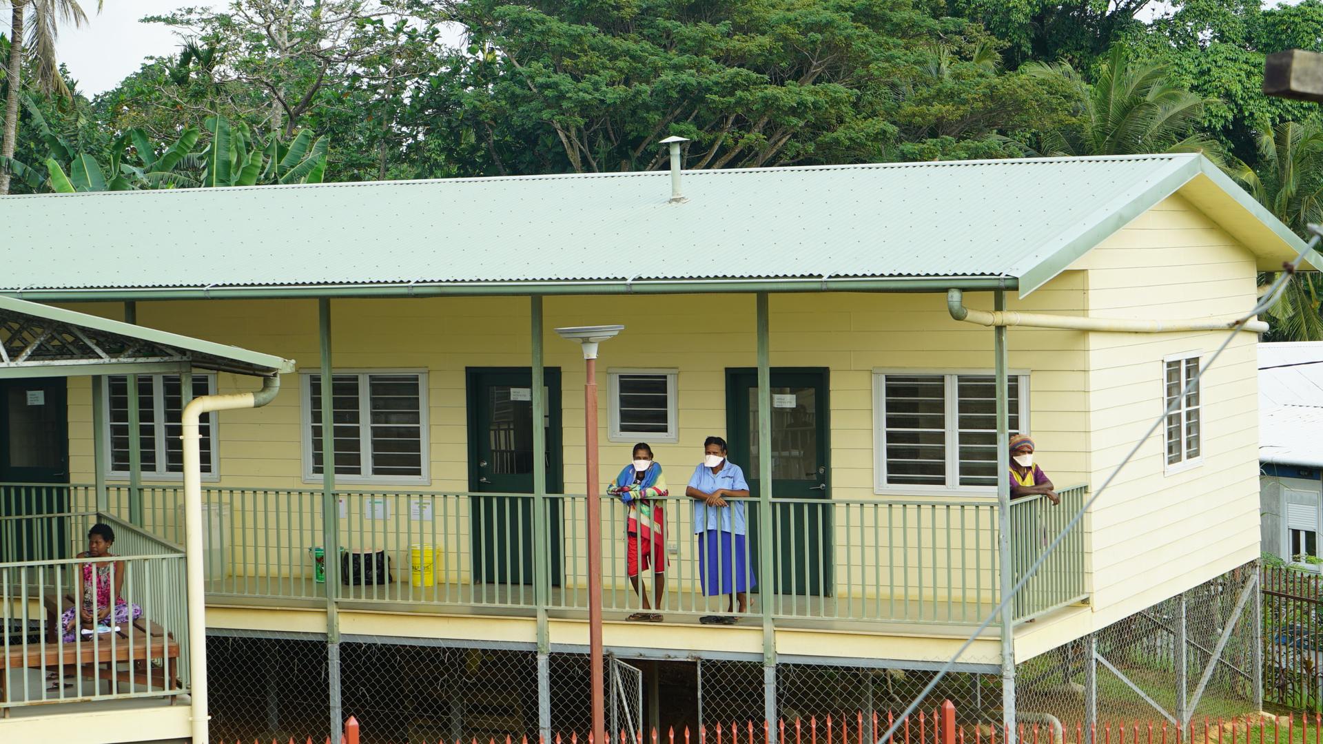 People stand with face masks on the verandah of a yellow hospital building. 