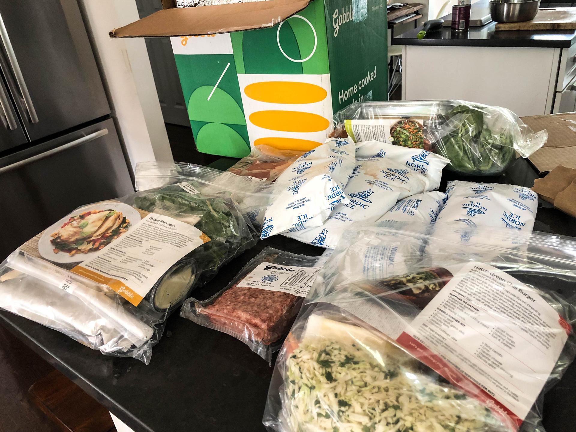 A meal kit is unboxed on a counter space. Food is wrapped in plastic packaging.