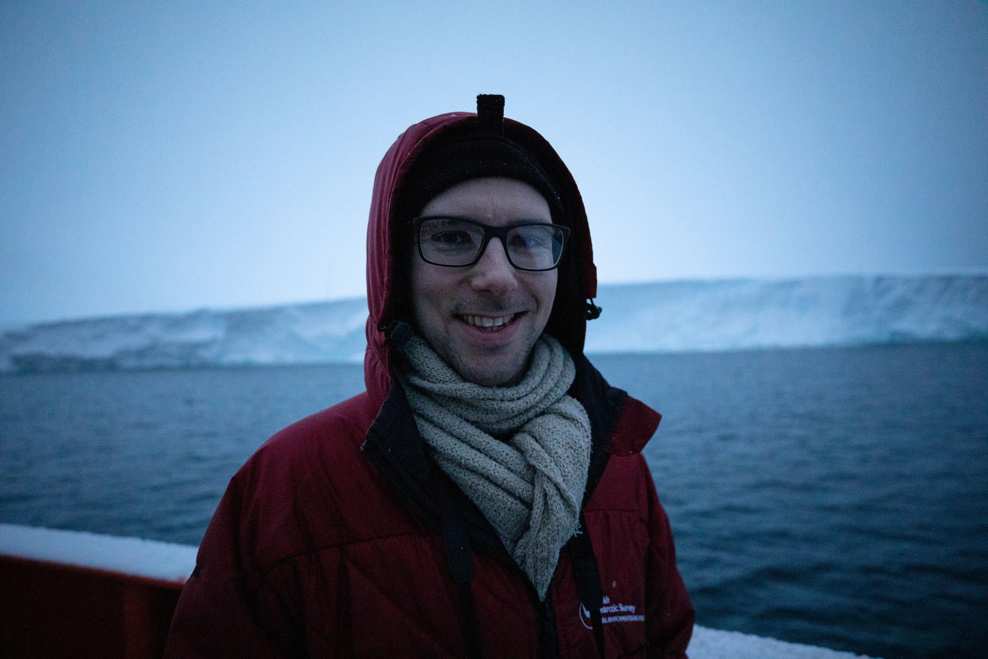 Oceanographer Peter Sheehan is shown with a red jacket and white scarf with Thwaites glacier in the background.