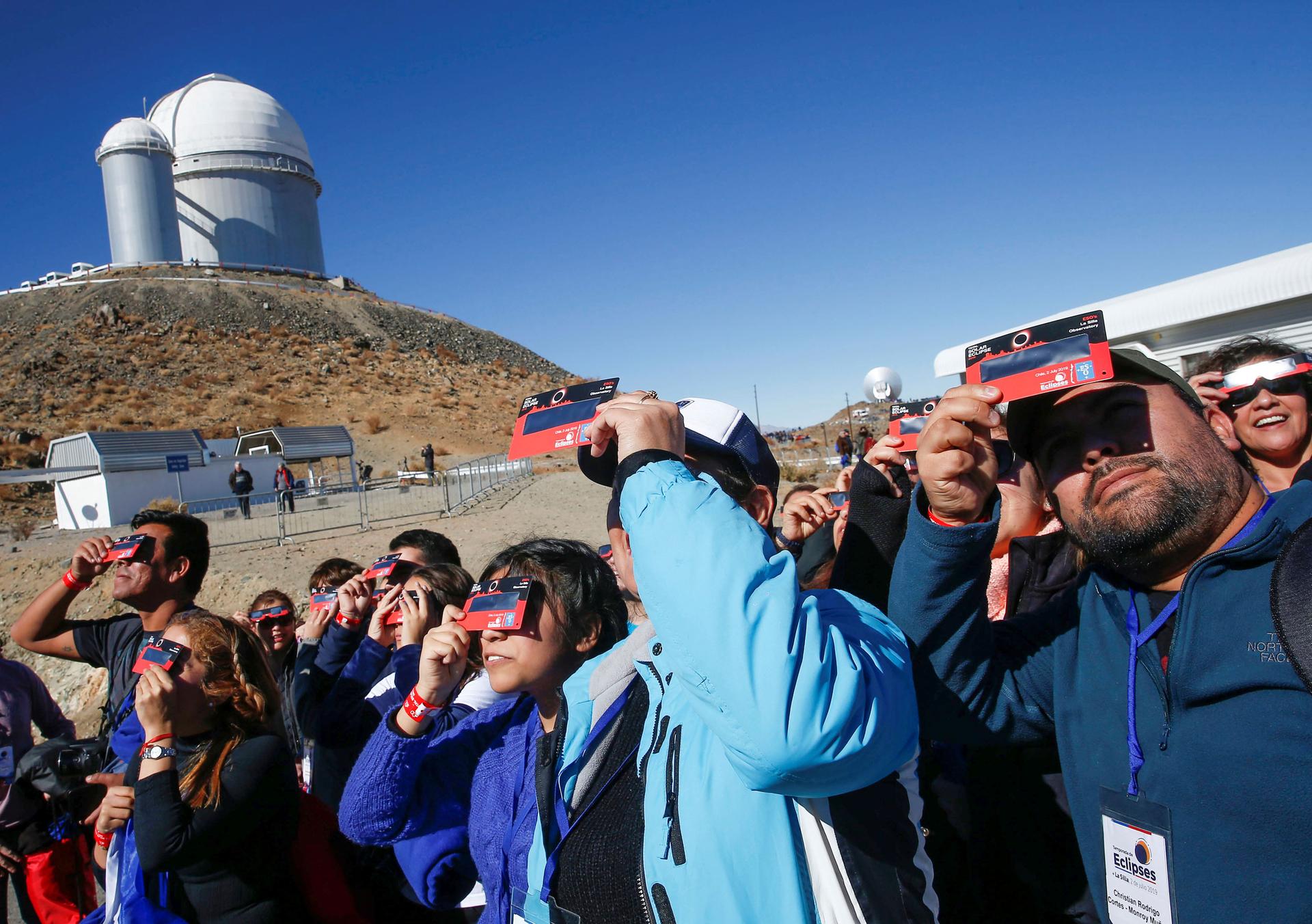 A large group of people are shown looking up at the sky with a paper solar eclipse viewfinders.