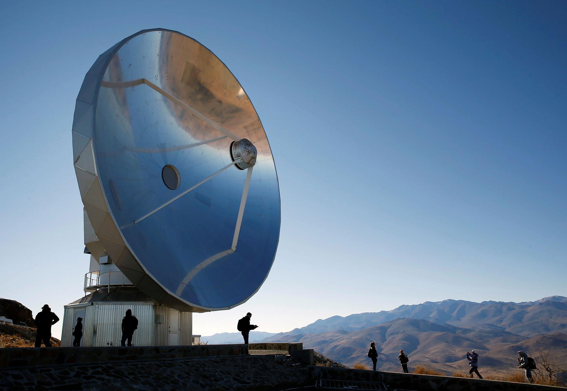 A large telescope is shown shining in the sun with people underneath.