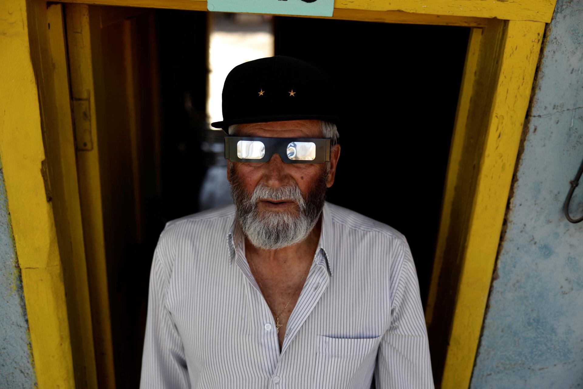 A portrait photograph shows an older man with shiny solar eclipse glasses standing in a yellow door frame.