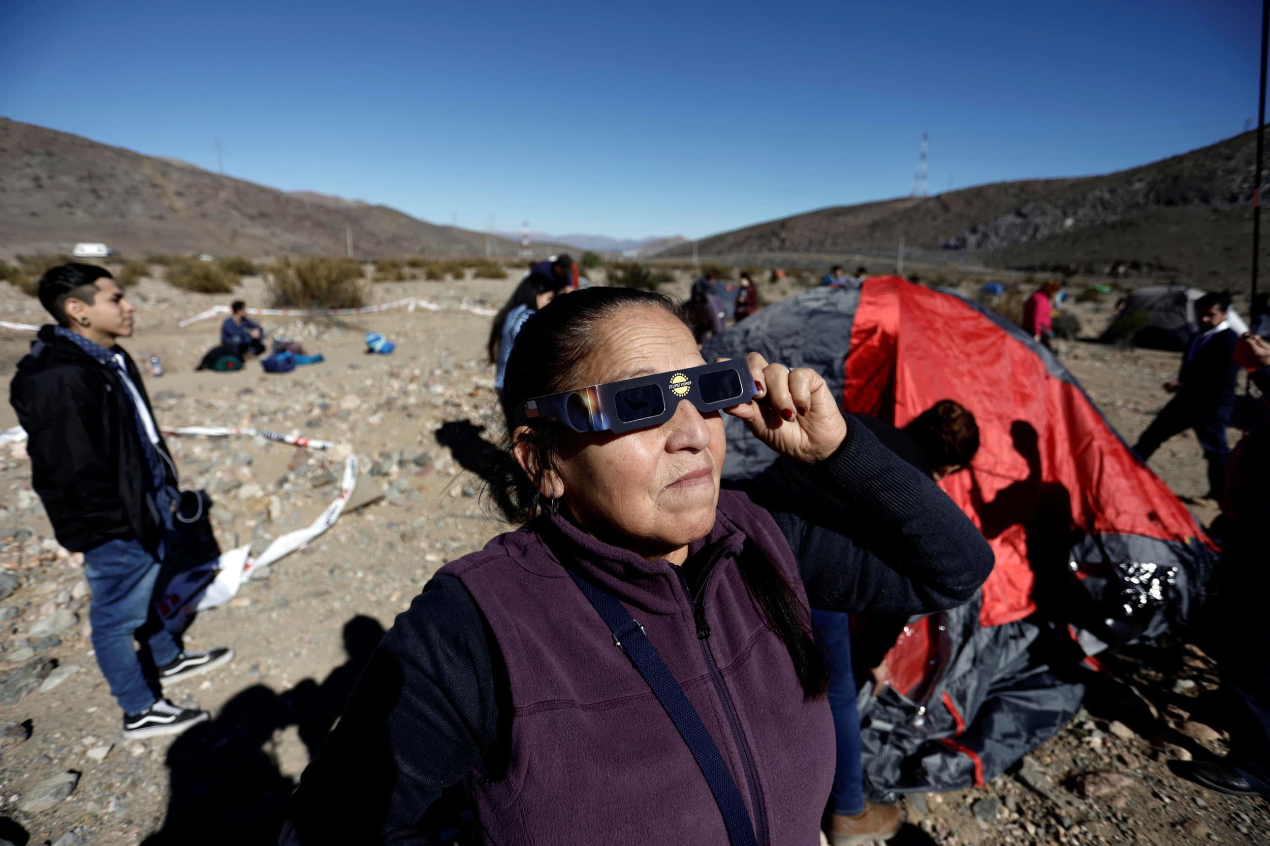 A woman wearing a purple vest is shown holding solar eclipse glasses to her face.