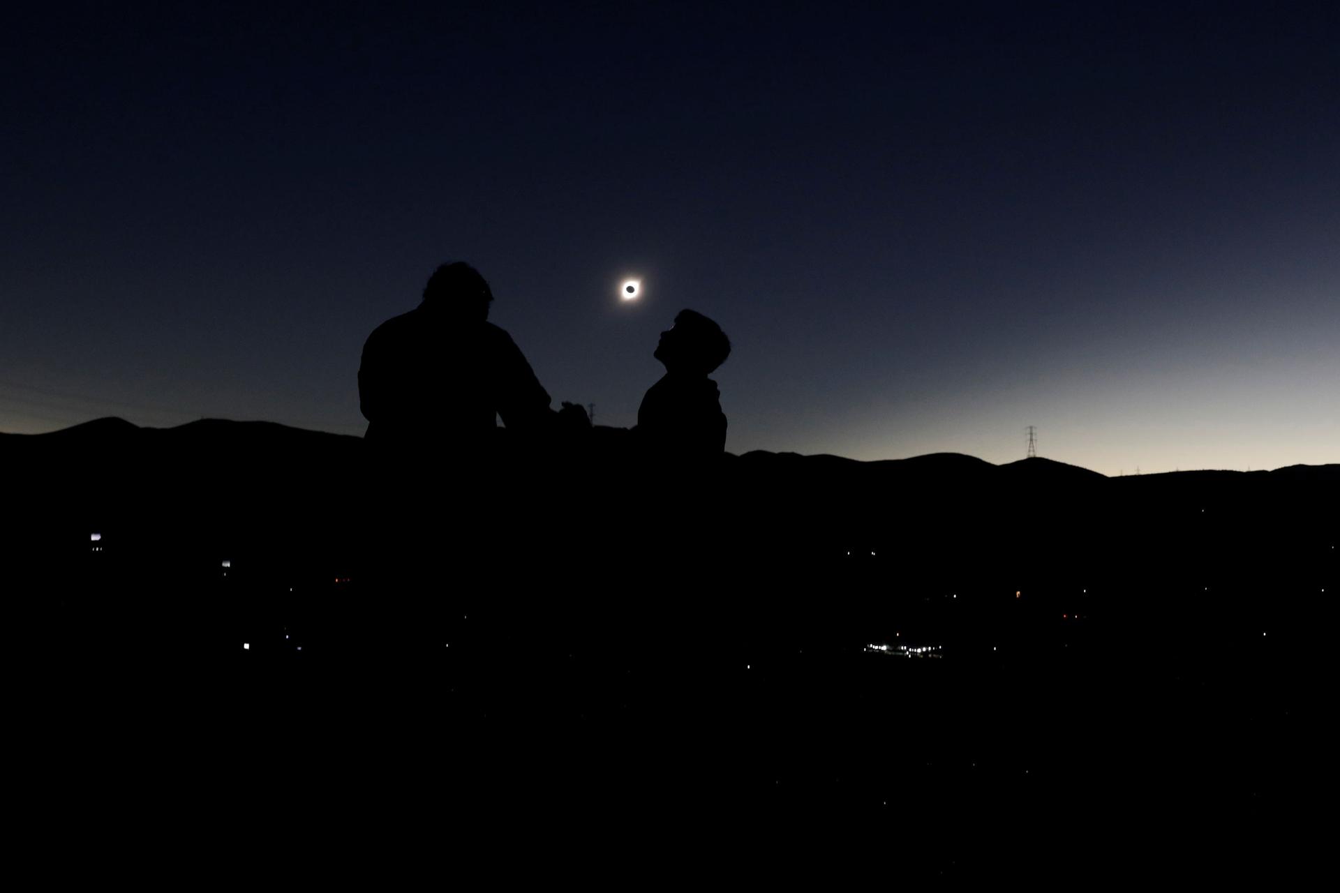 A solar eclipse is shown in the distance framed between two people in the nearground in shadow.