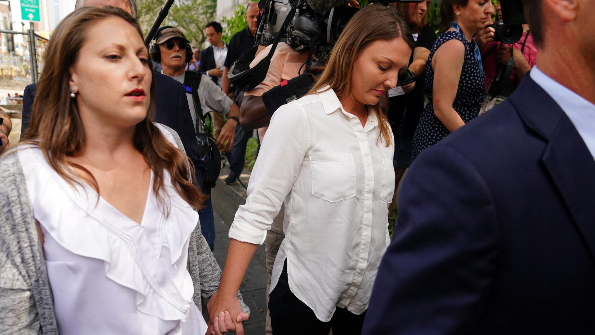 Two women walk with their hands clasped together as they're surrounded by a crowd of reporters