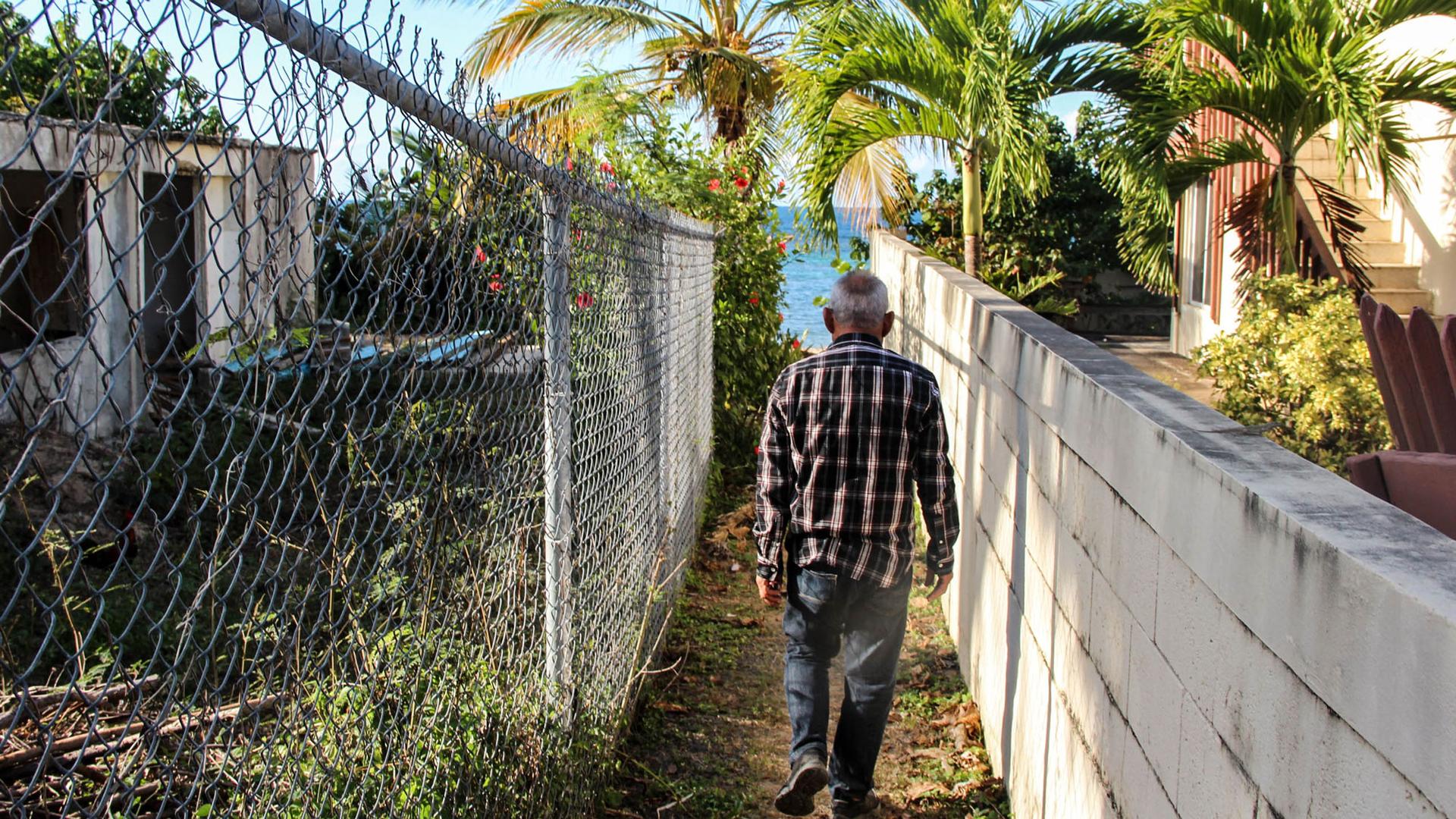 A man is shown walking between two houses with a chain link fence on his left and a manicured stone wall on his right.