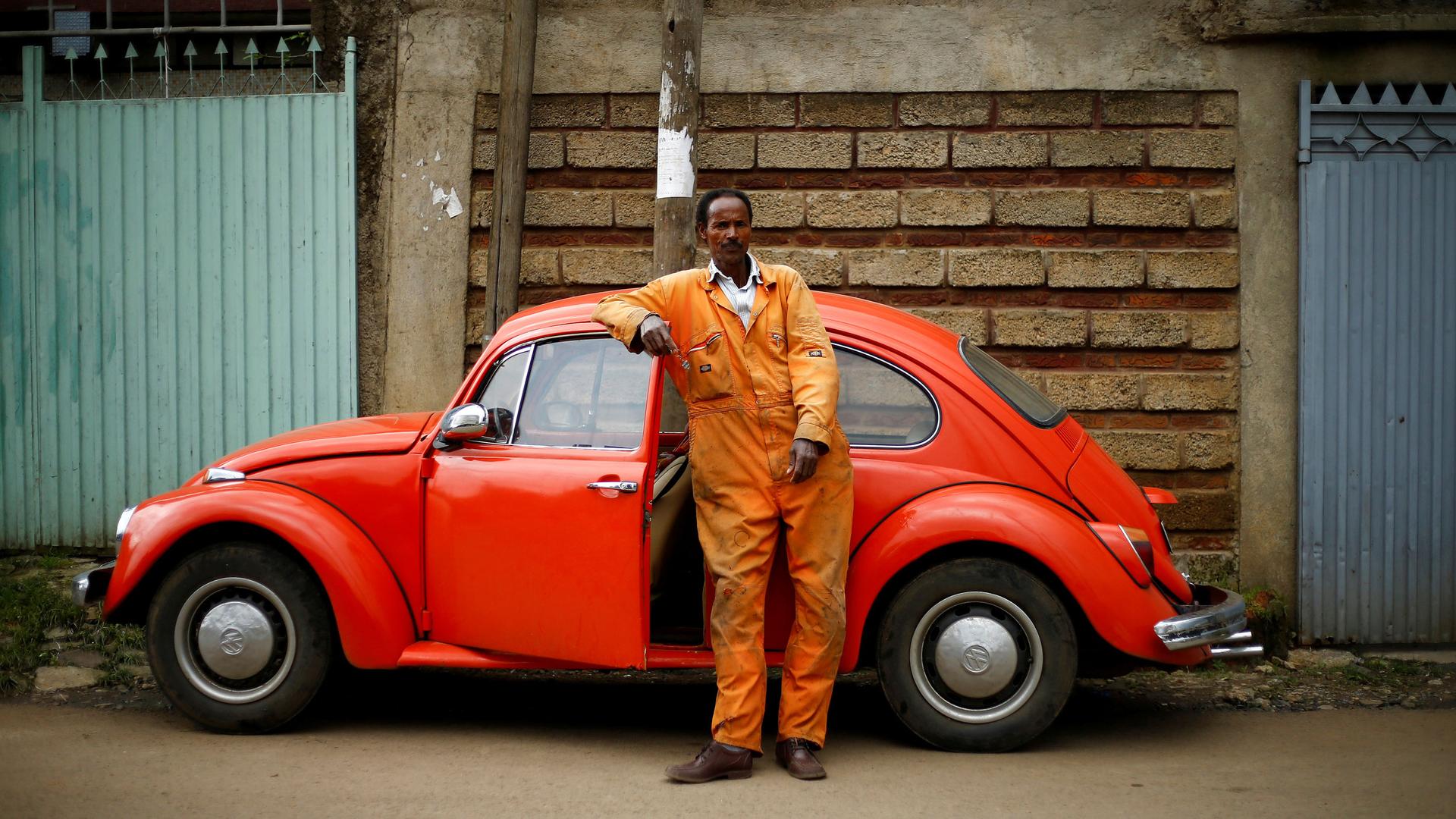 A man in an orange jump suit leans his arm on a red 1965 model Volkswagen Beetle car.
