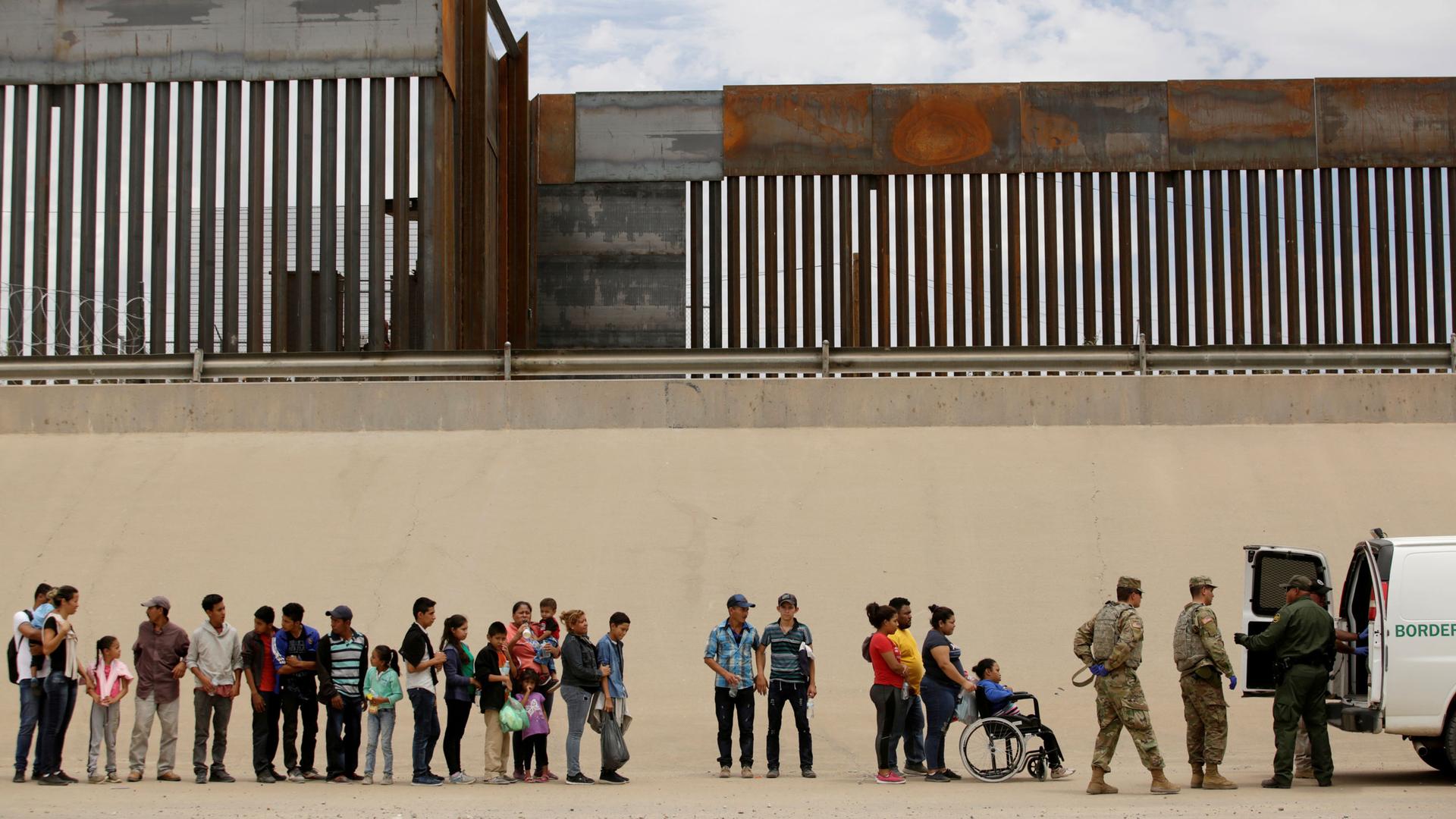 A line of migrants that includes young and older people along with a person in a wheelchair are shown standing next to the large border fence.