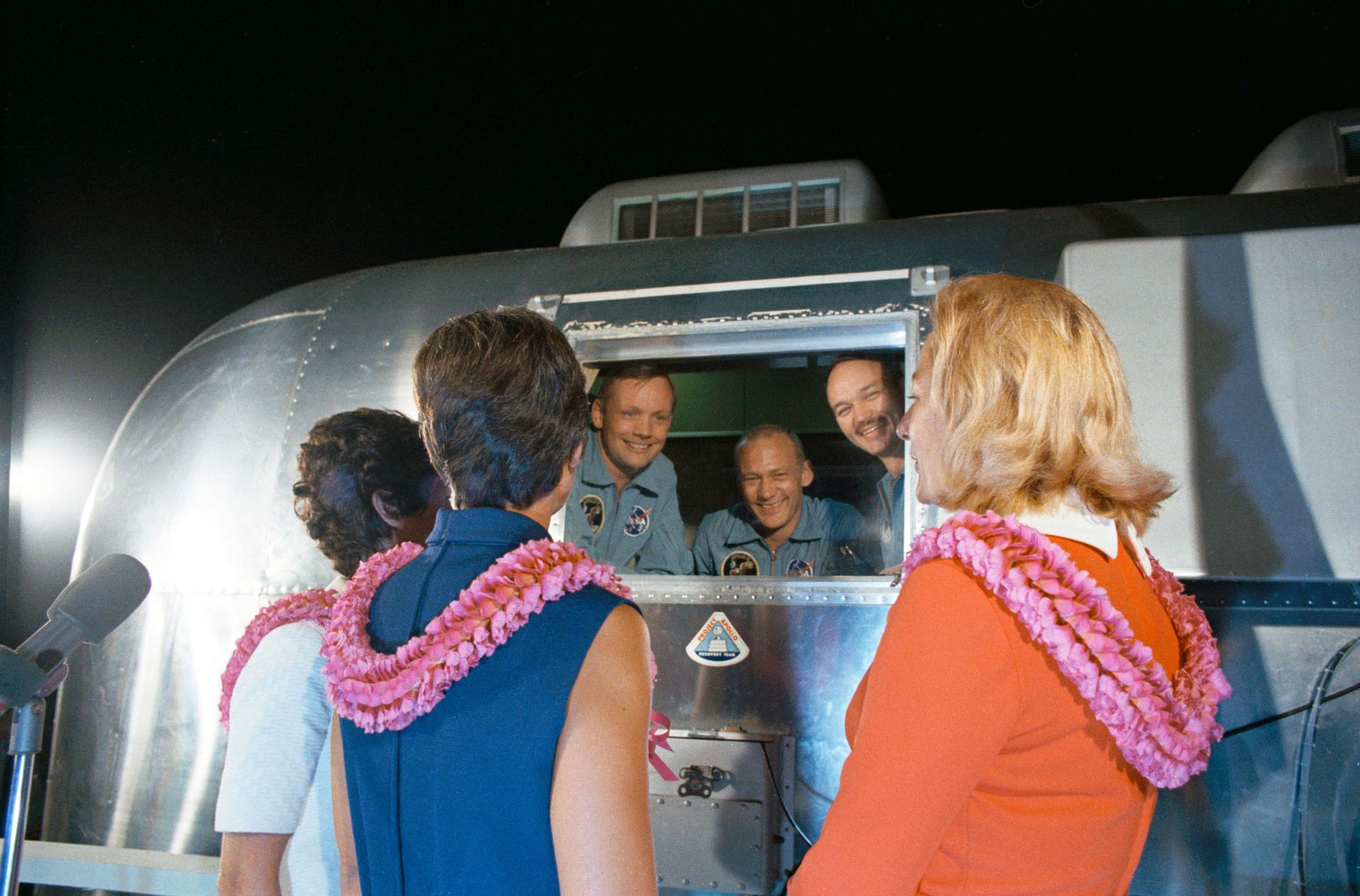 A silver-colored containment structure is shown Neil A. Armstrong, Edwin E. Aldrin Jr., and Michael Collins speaking with their wives.
