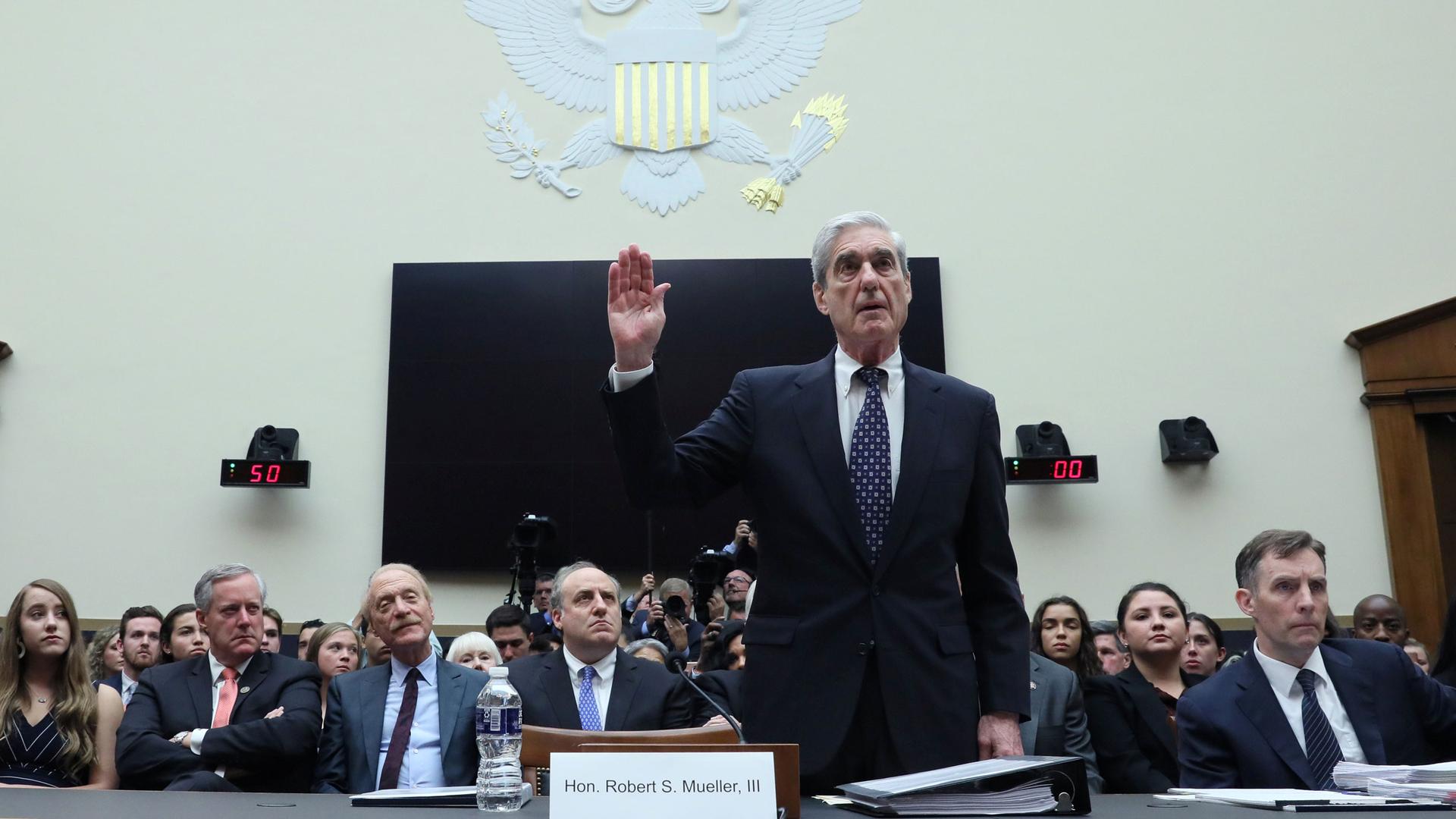 Former Special Counsel Robert Mueller is shown with his right hand raised with a crowd of people seated behind him.