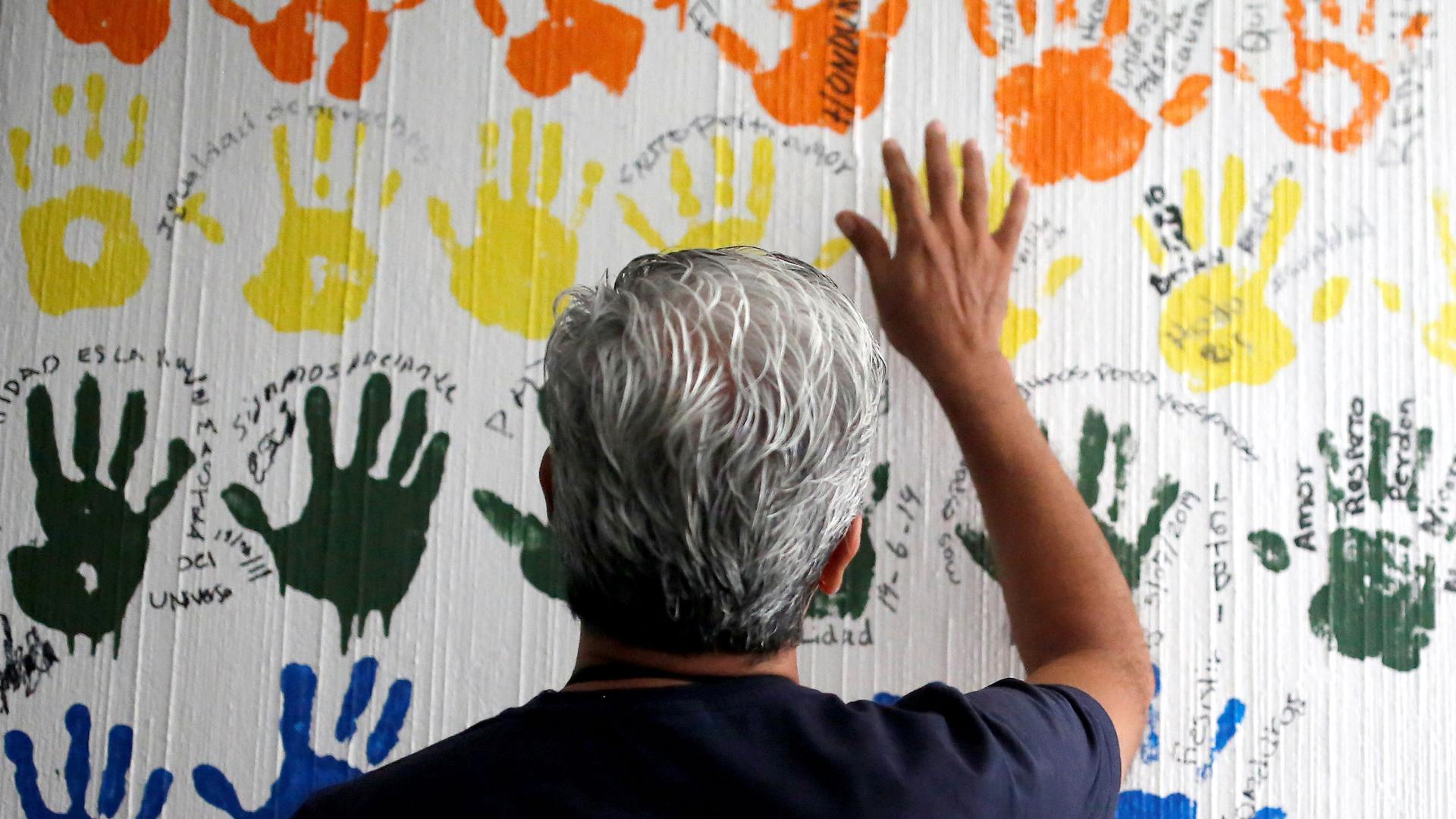 a man holds up his right hand in front of a wall of fingerpaint-hands
