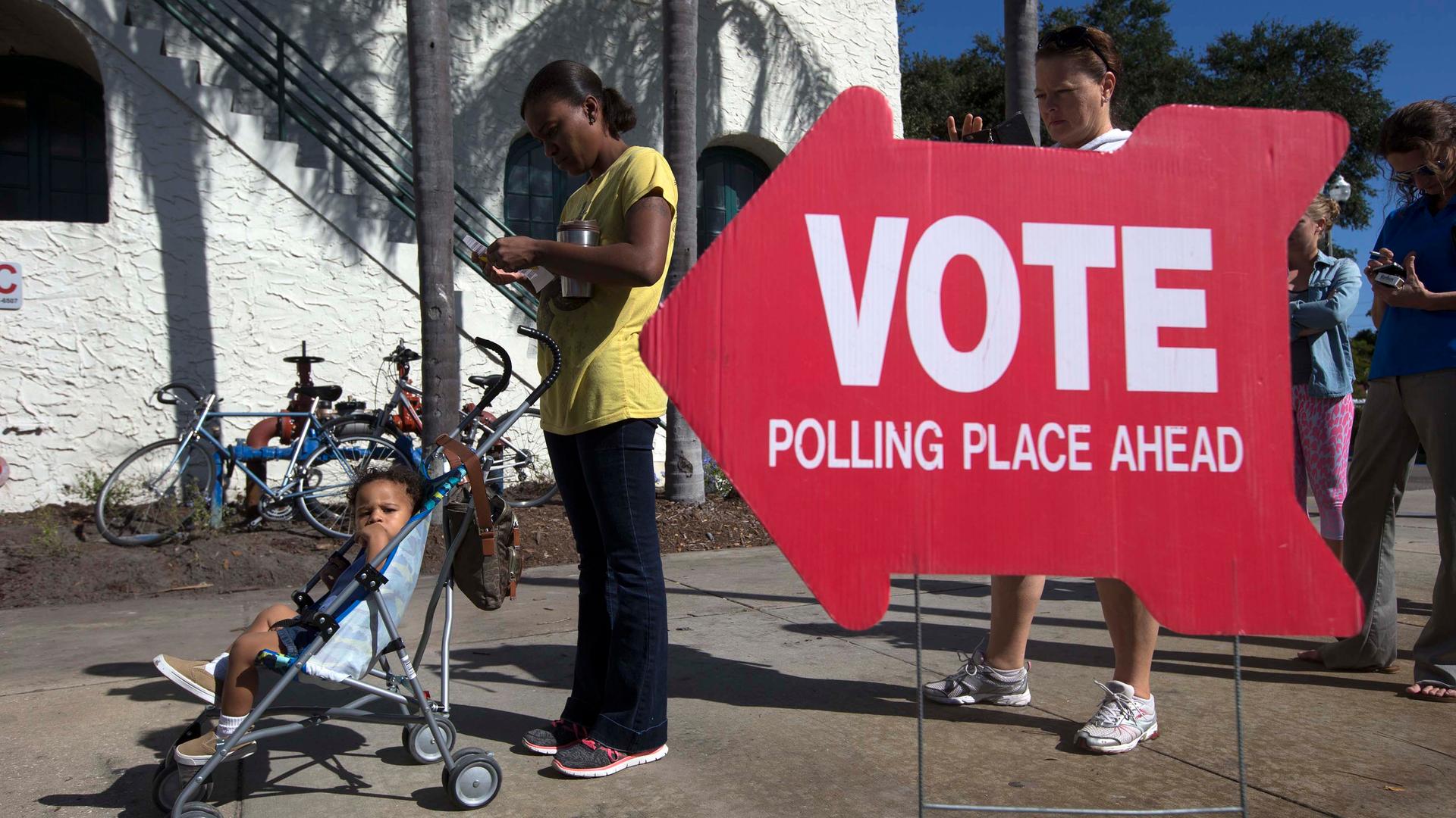 A sign saying vote in the foreground with people and a child in a stroller in the foreground