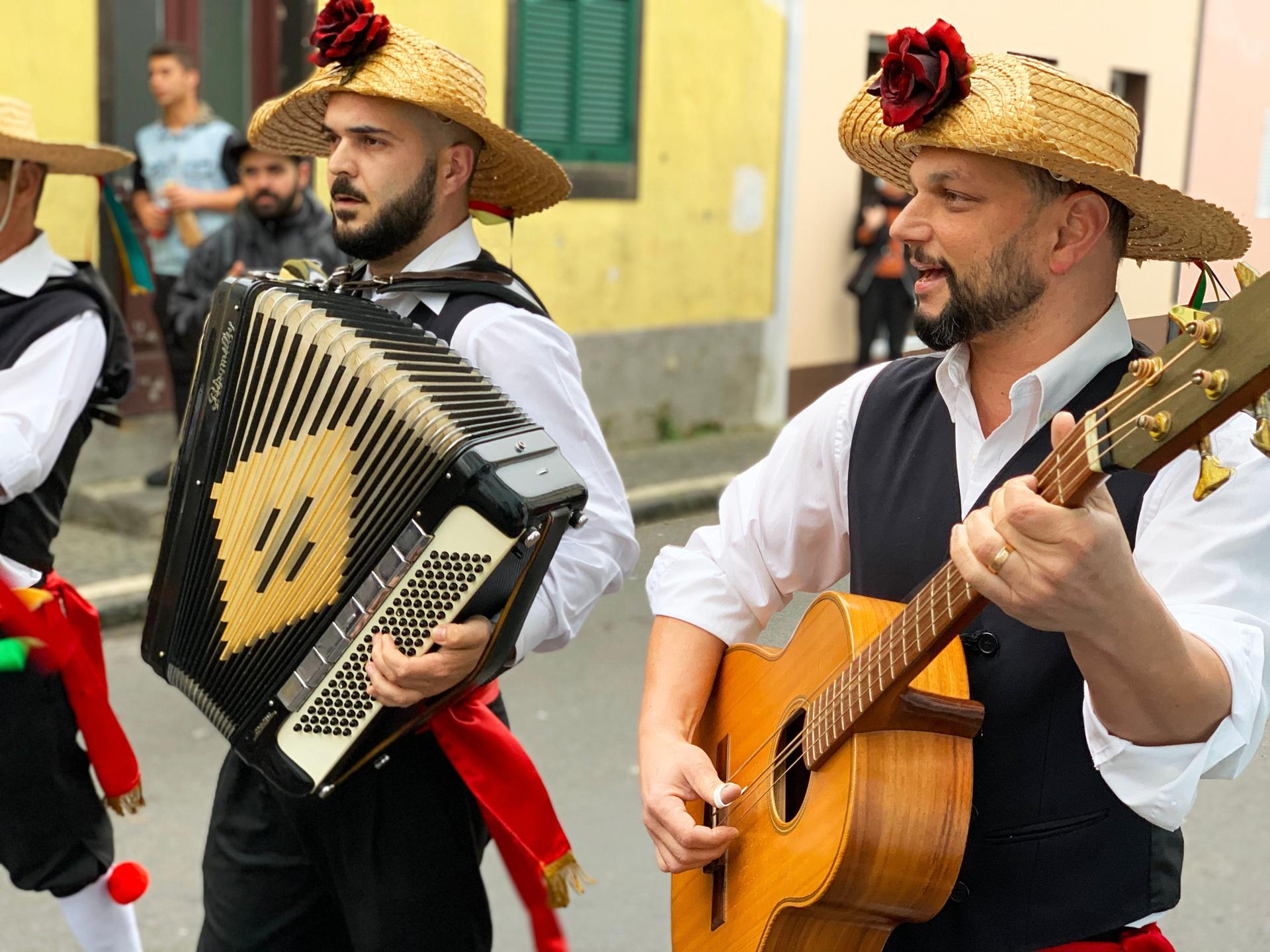 Musicians walk down the street in hats. One plays a guitar, one an accordion.