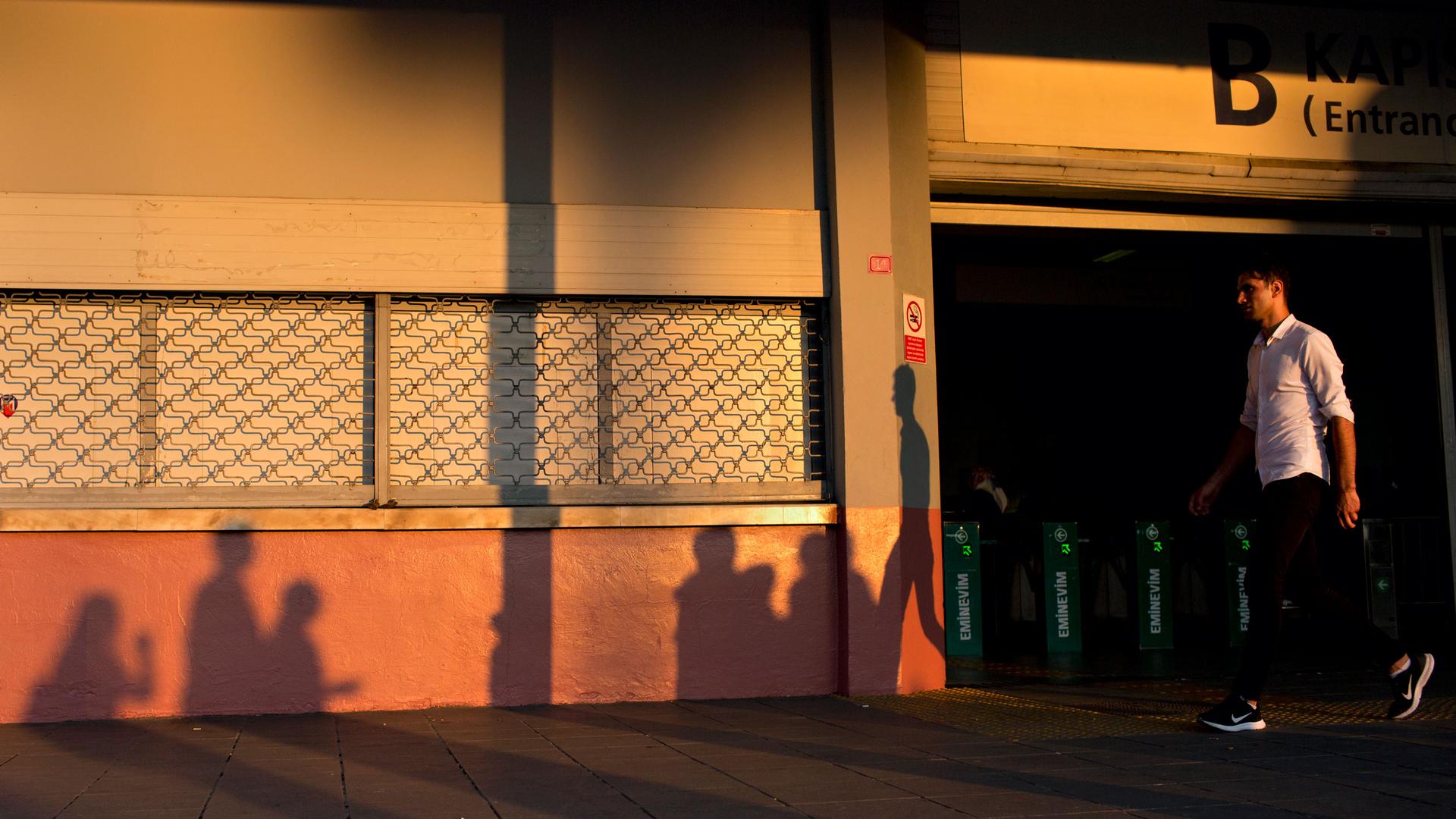 A man is shown walking in front of a metro stop in Istanbul at dusk with the long shadow of several other people on the wall.