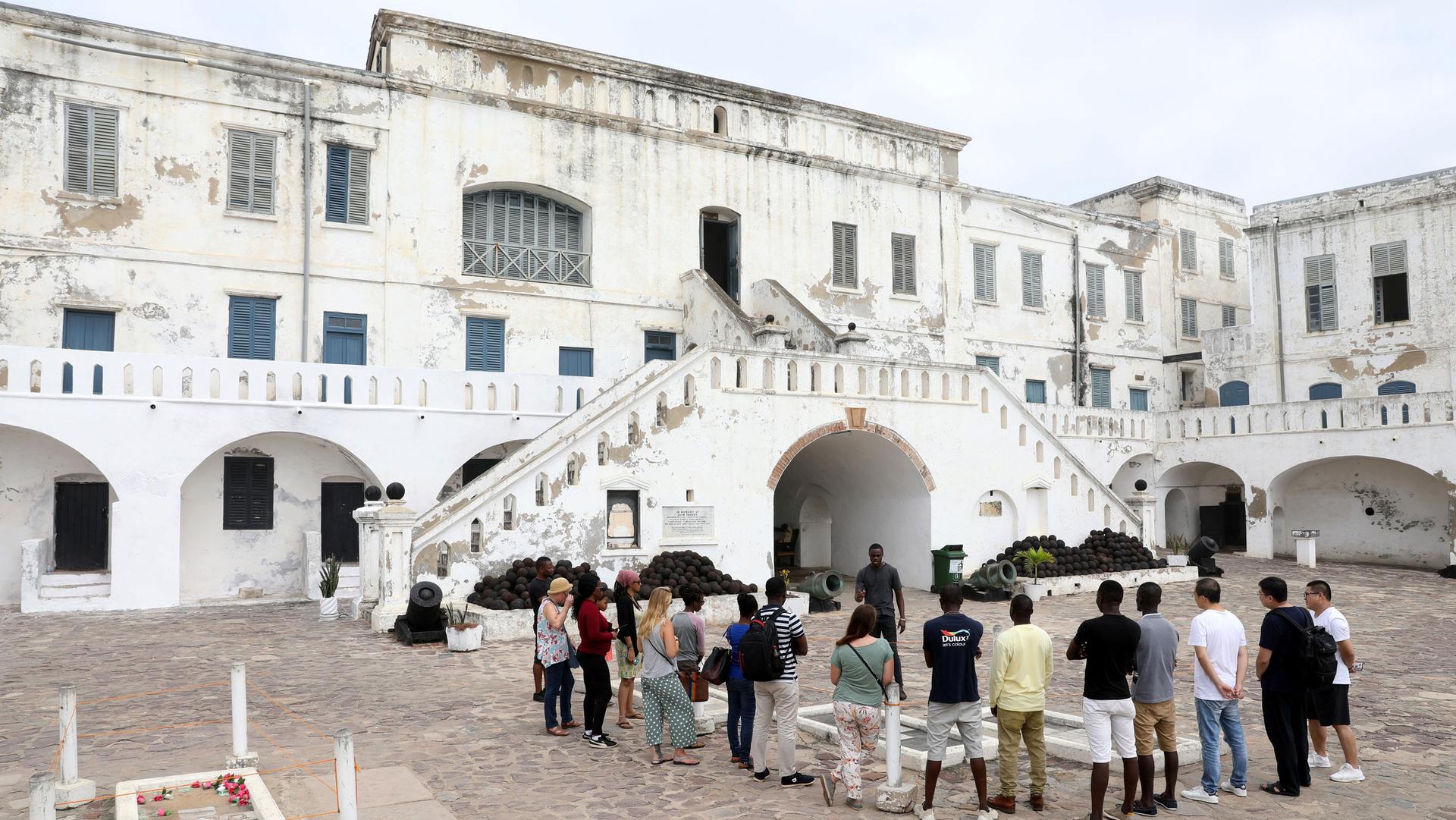 Several people are shown standing around a tour guide in front of the white facade of the Cape Coast Castle.
