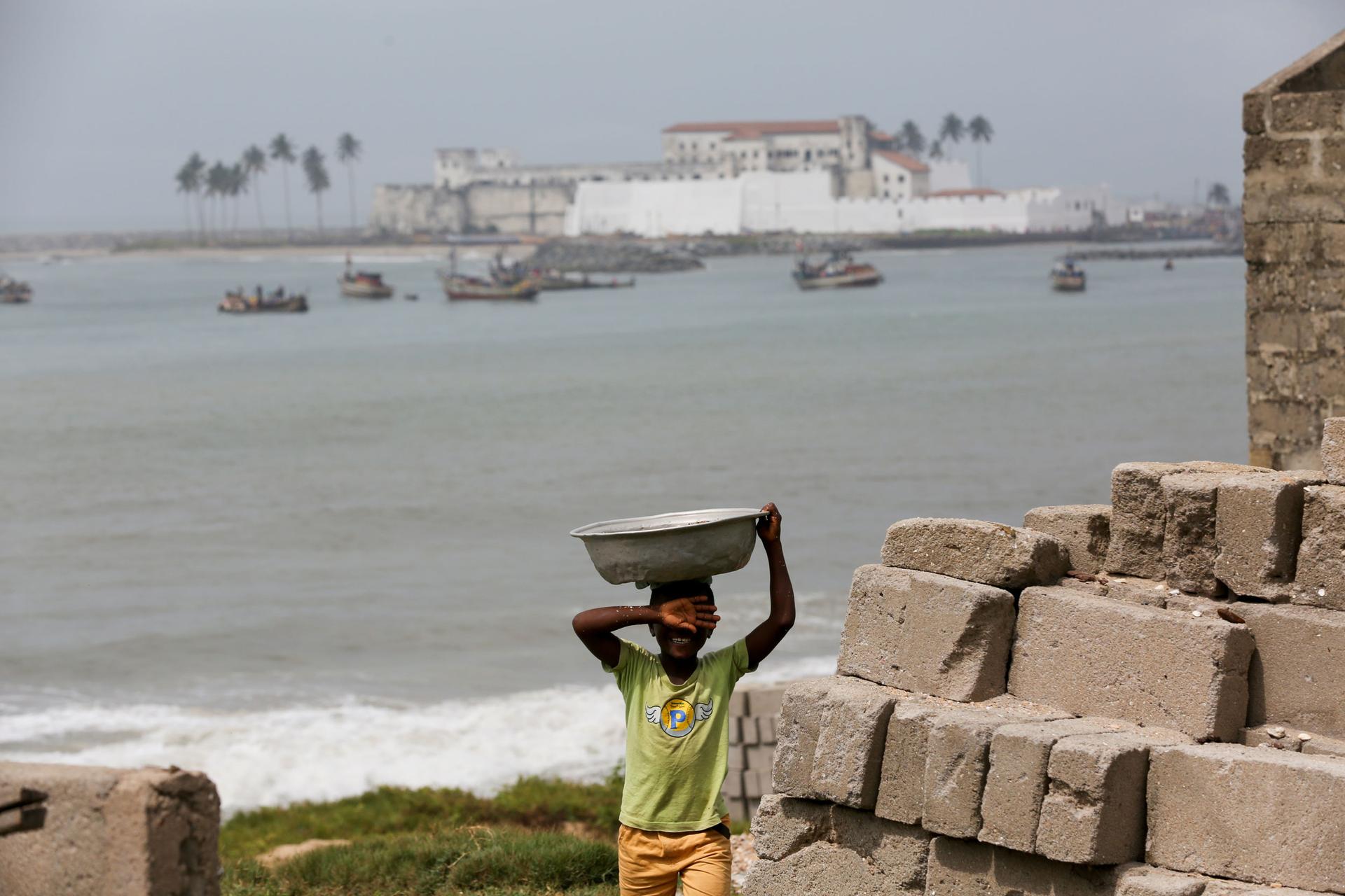 A young boy is shown in the nearground holding a large pan on his head with the ocean and Elmin Castle in the background.