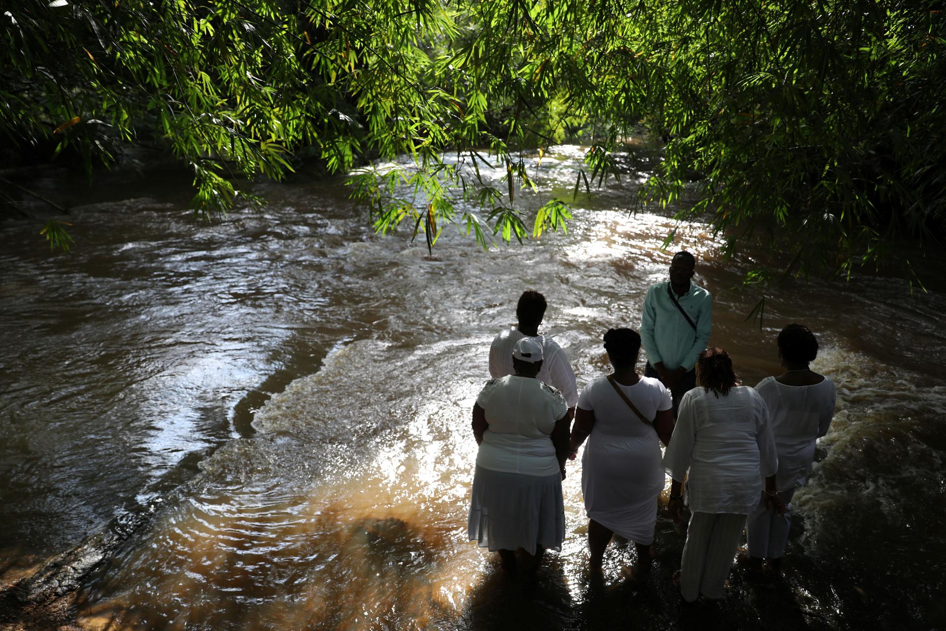 A group of people are shown wearing white and standing ankle deep in a river.
