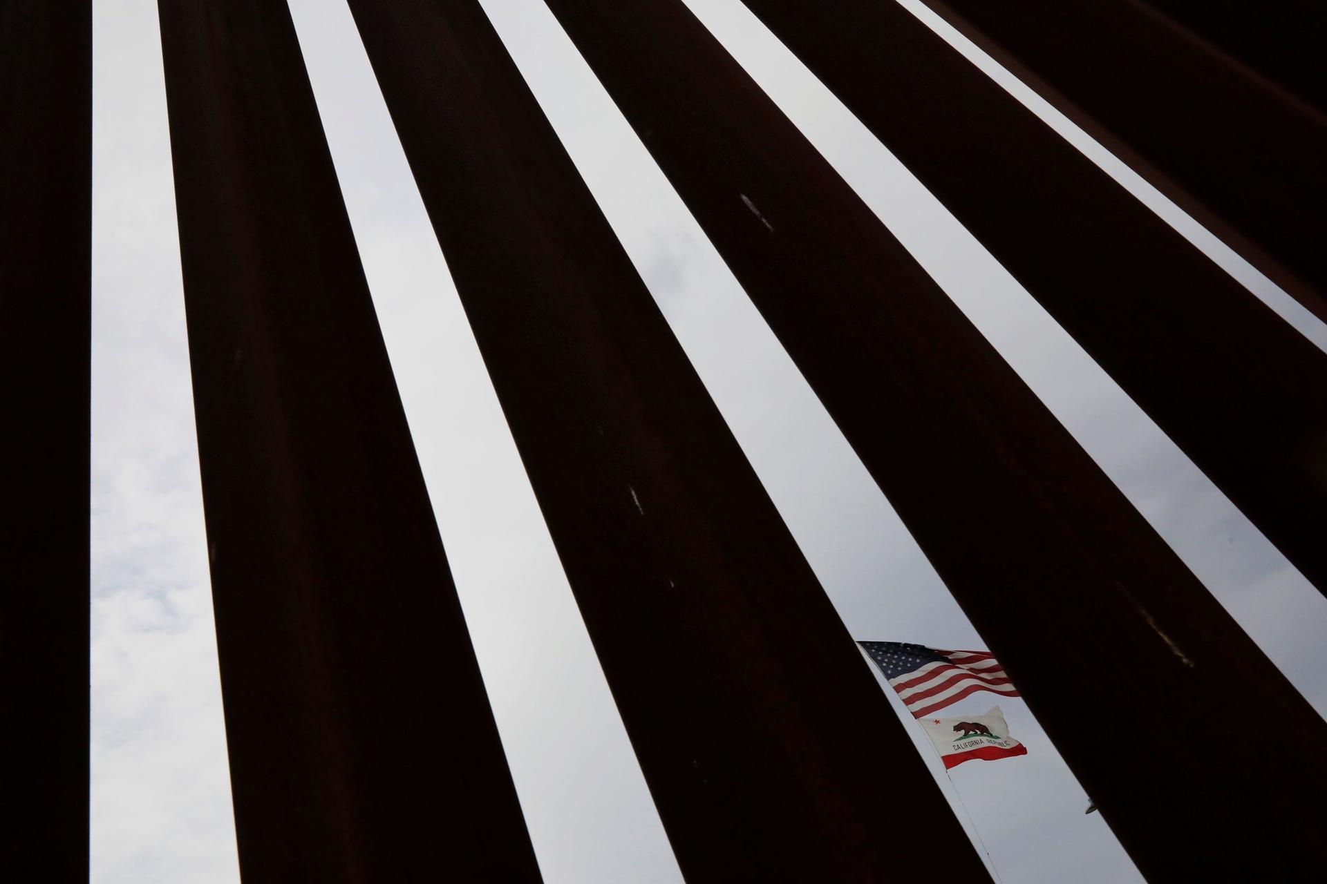 A photograph with dark metal slats are shown in contrast to the blue sky in-between with the US and California flags in one of the slots.