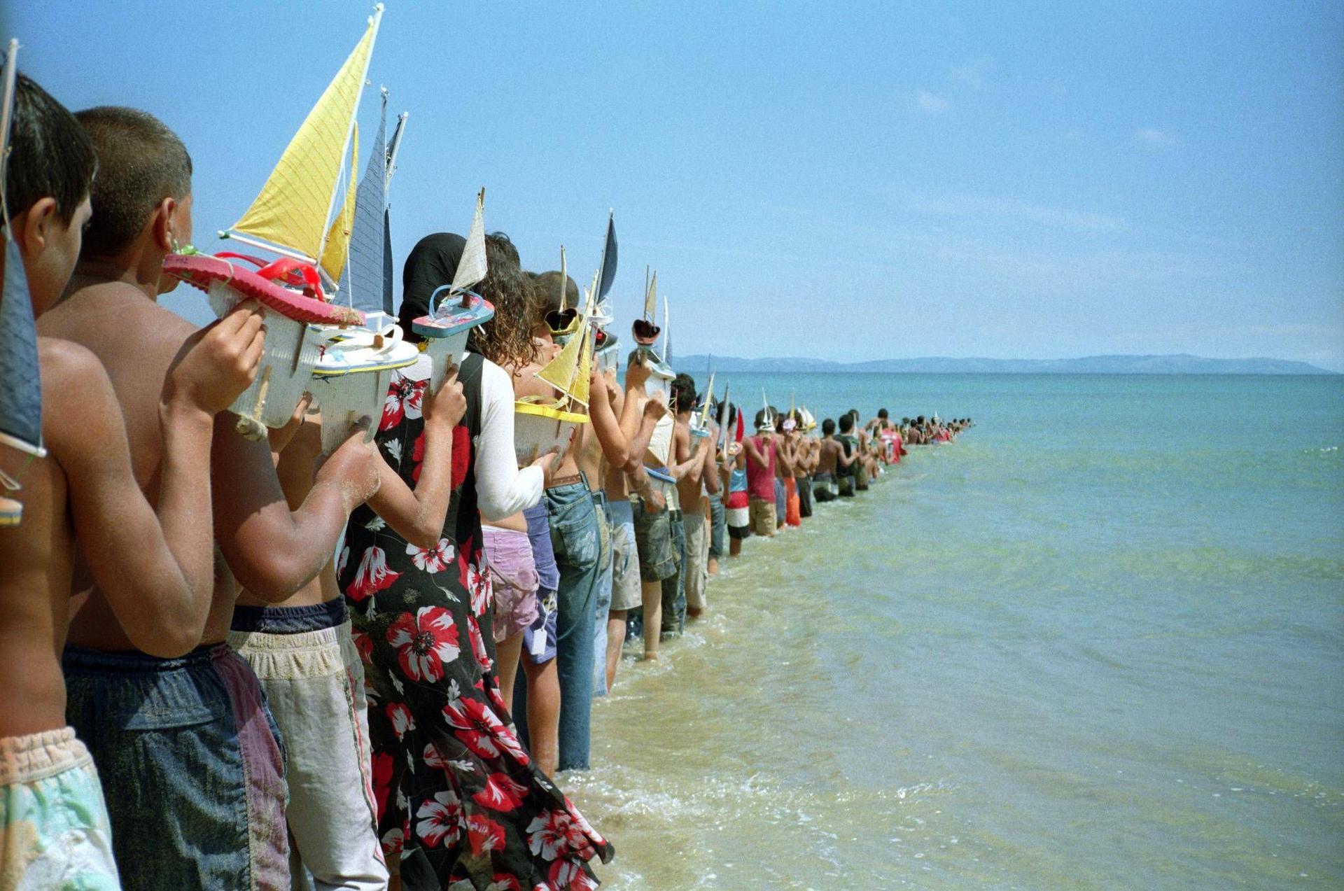 A line of people holding toy boats is seen going into the ocean.