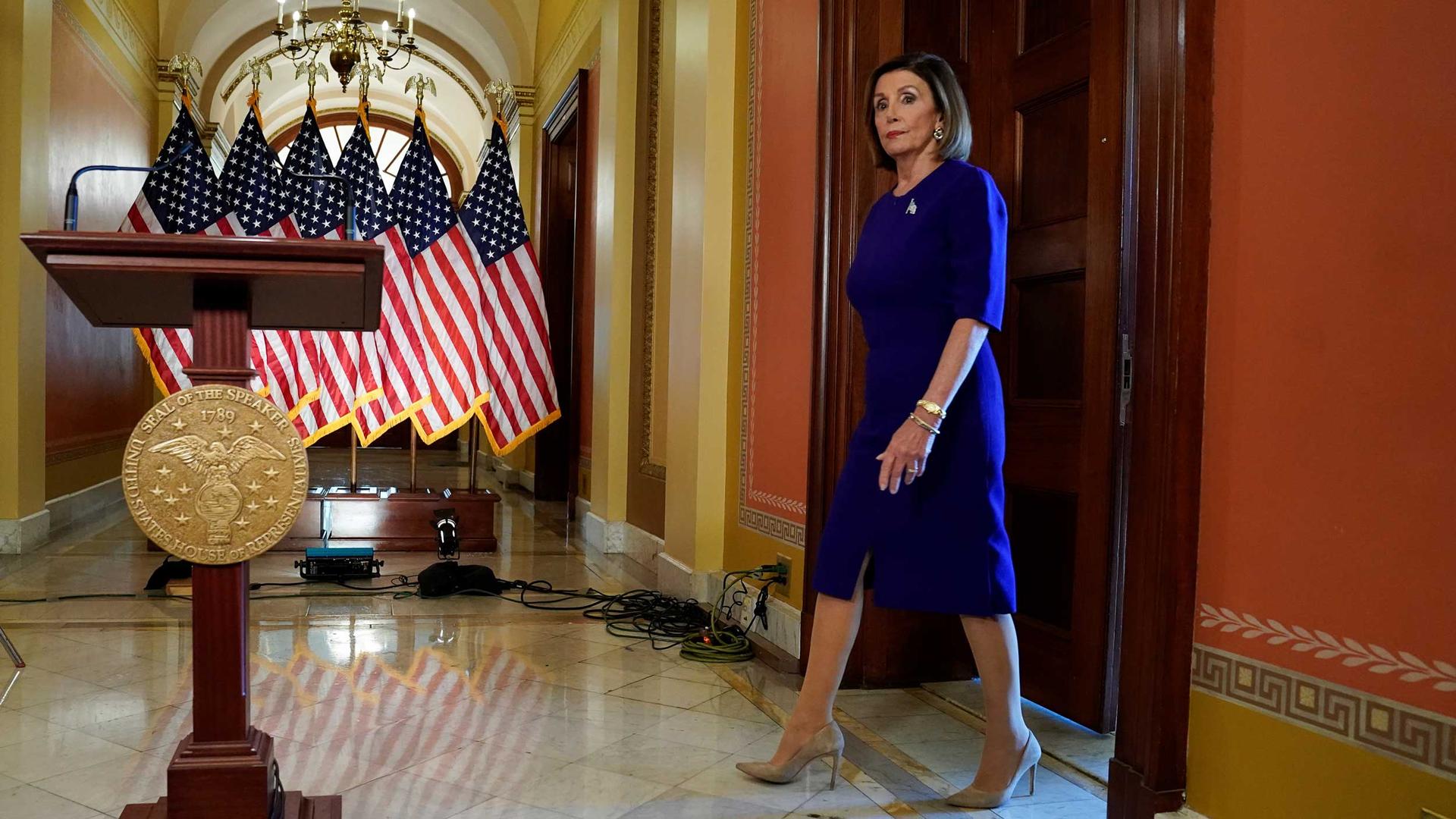 Nancy Pelosi walks up to a podium with a line of American flags set up behind it