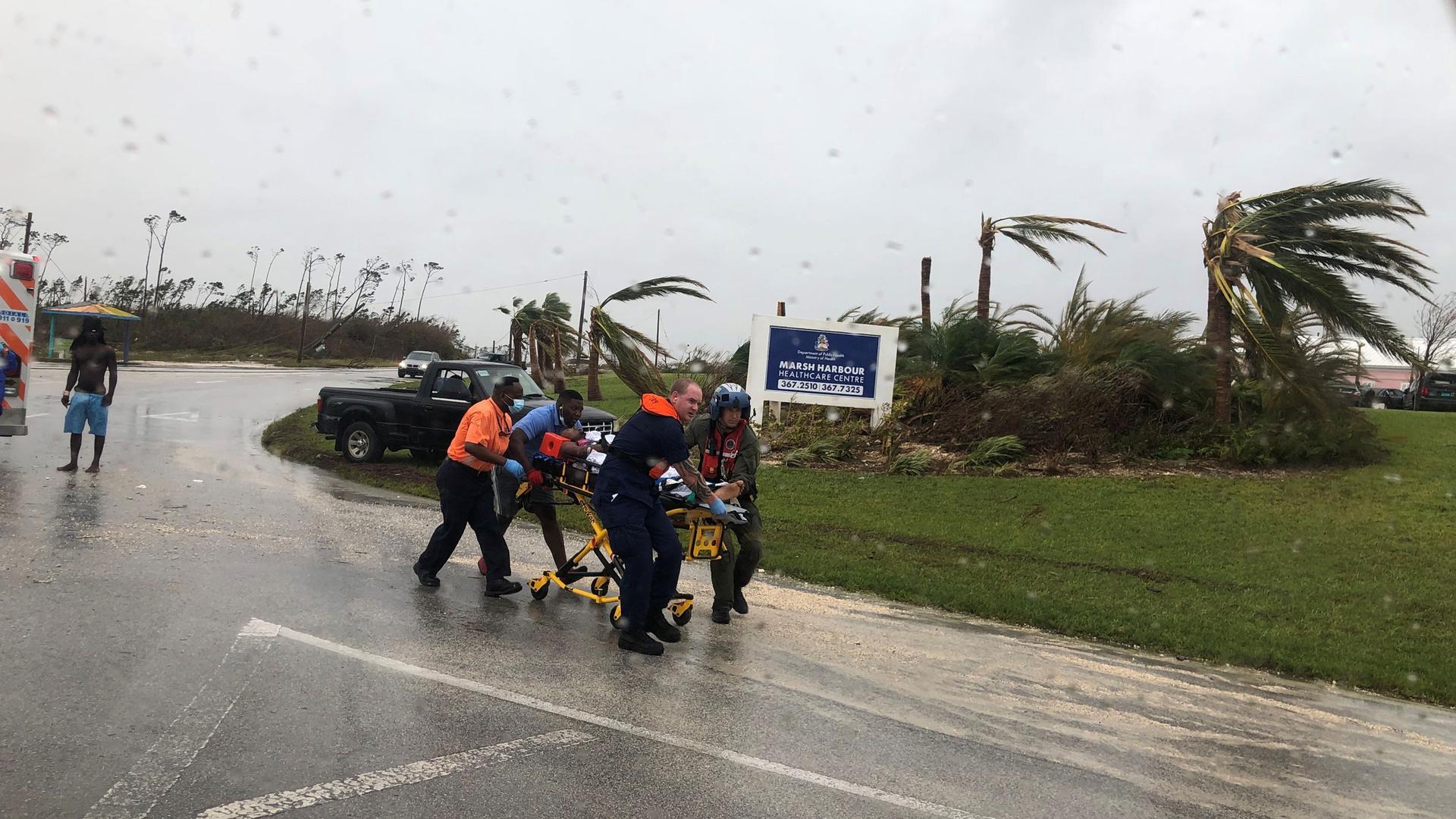 Several rescue workers are shown moving a yellow emergency stretcher down a road with winds blowing the trees in the background.