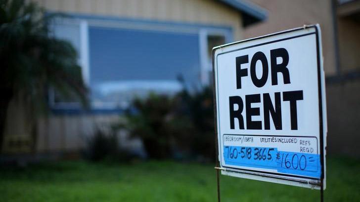 A "For Rent" sign in focus in font of a house. 