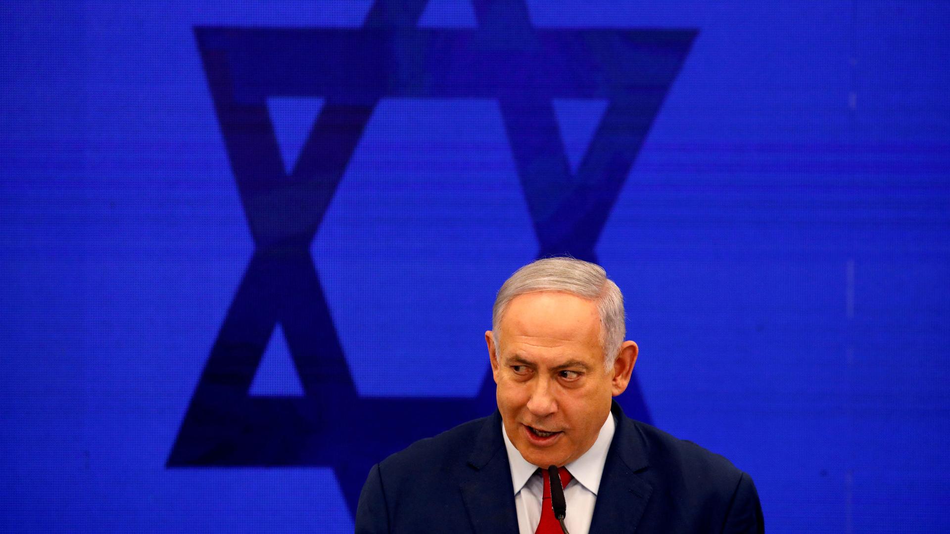 Israeli Prime Minister Benjamin Netanyahu is shown wearing a dark suit and red tie with the Star of David behind him.