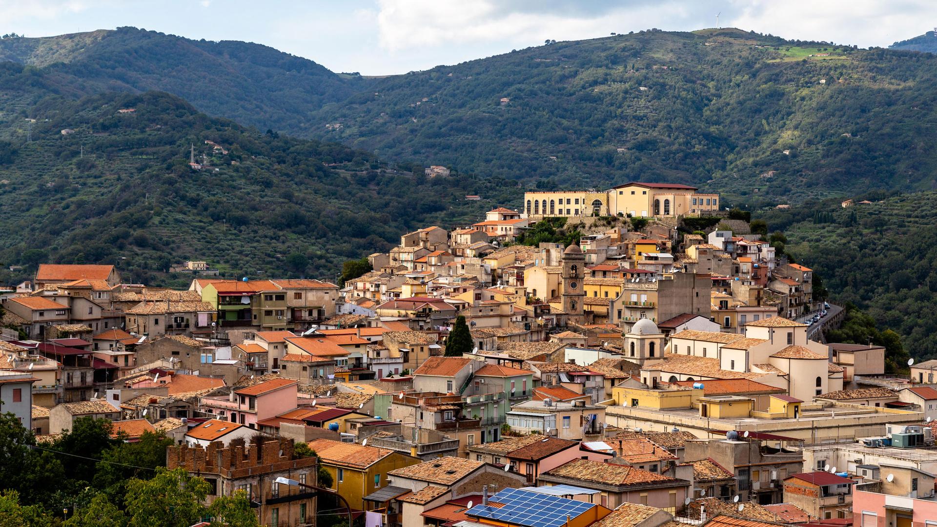 An old Italian town of San Piero Patti is shown from above with the rooftops of homes built into the lush green hillside.