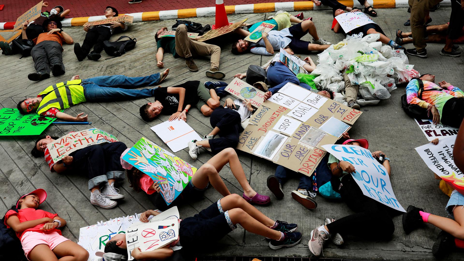 Several young people are shown laying down on the ground with placards protesting for climate change action.