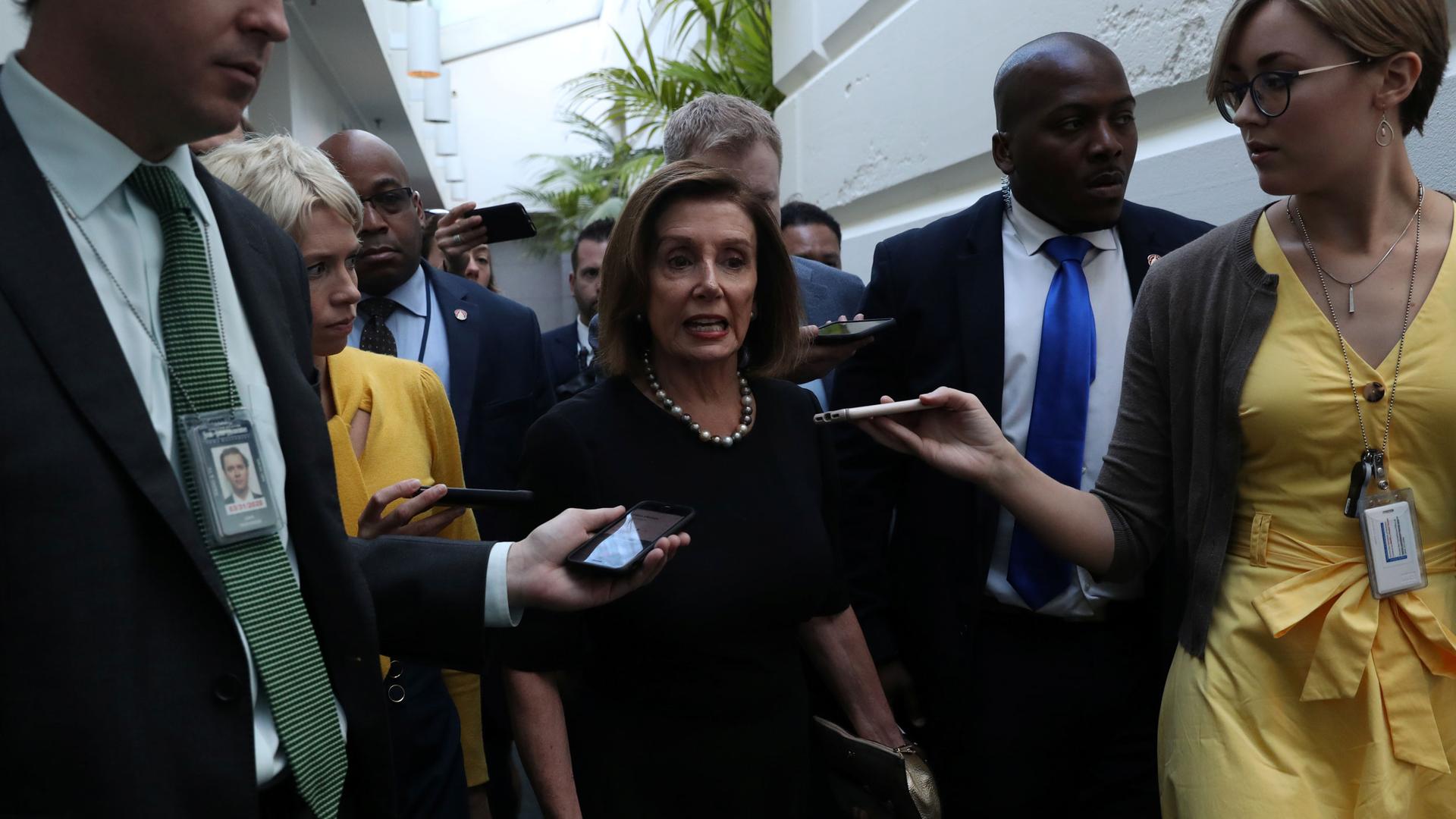 US House Speaker Nancy Pelosi is shown wearing a dark dress and pearls while surrounded by reporters.