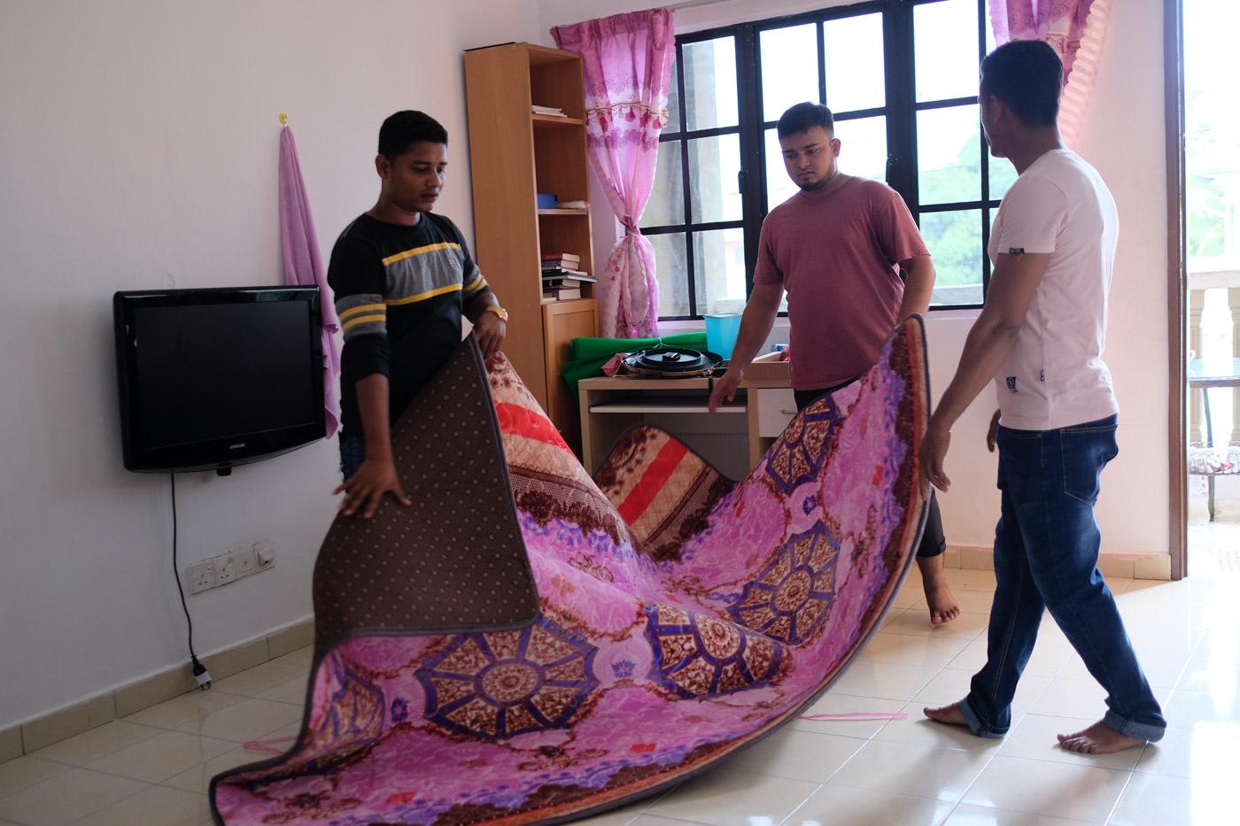 Three young men lay out a new, red and pink carpet in apartment