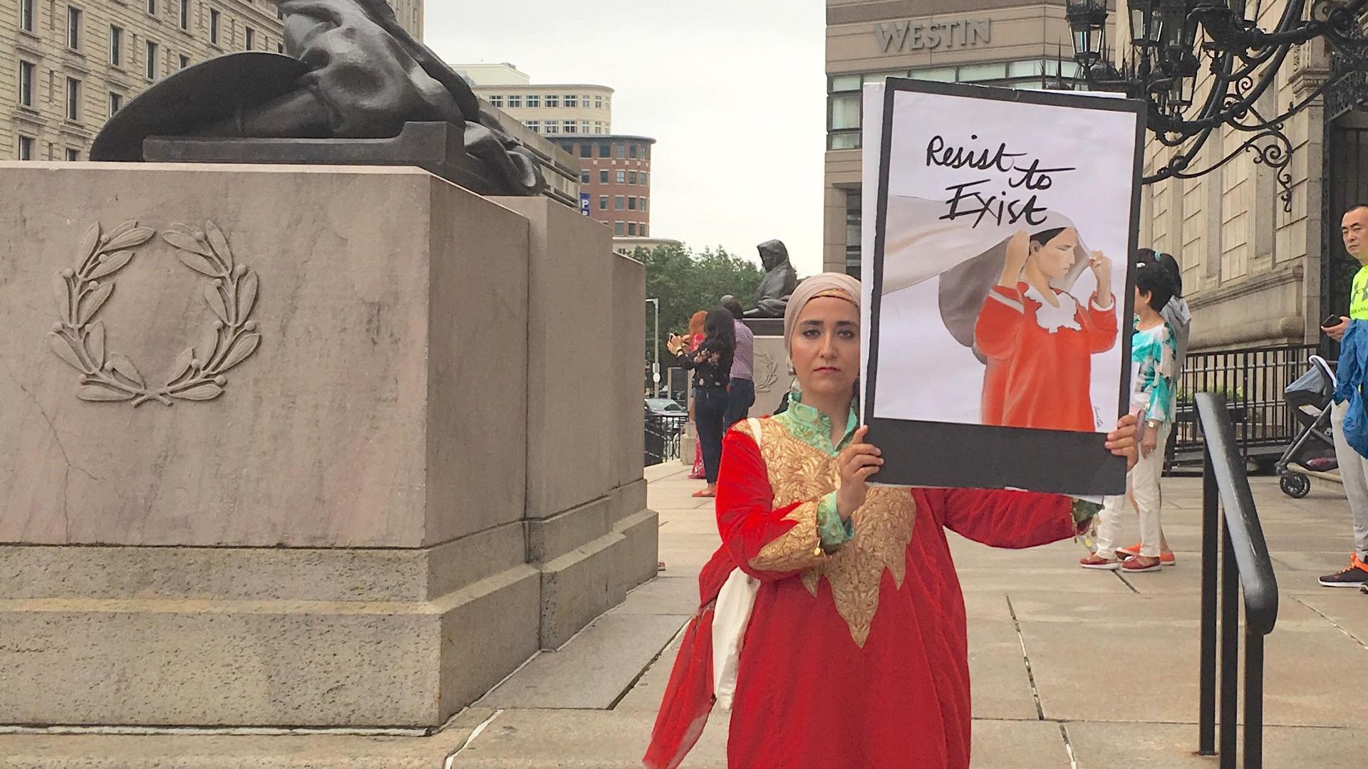 A woman in a red pheran stands holding an image of a woman in a red pheran