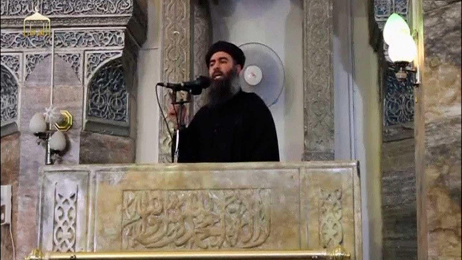 A man speaks at a podium. Behind him is the elaborately decorated wall of an interior of a mosque.