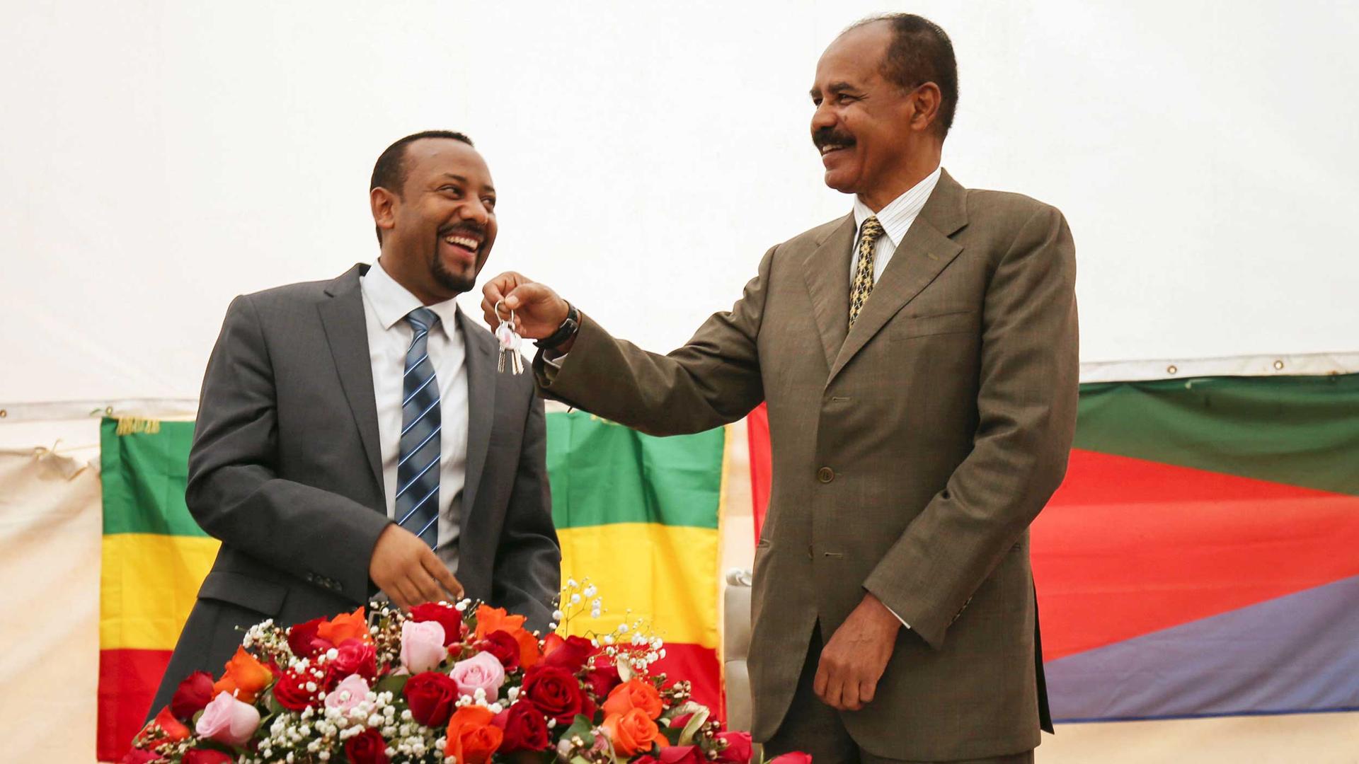 A man smiles as another man dangles keys from his hand. Behind the pair are the flags of Ethiopia and Eritrea.