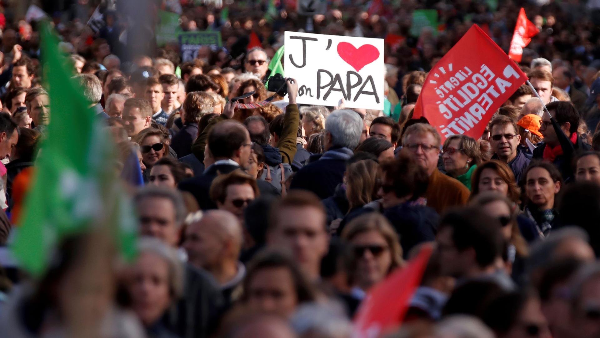 A crowd of people carry flags. One holds a sign that says "J'[HEART] Papa.