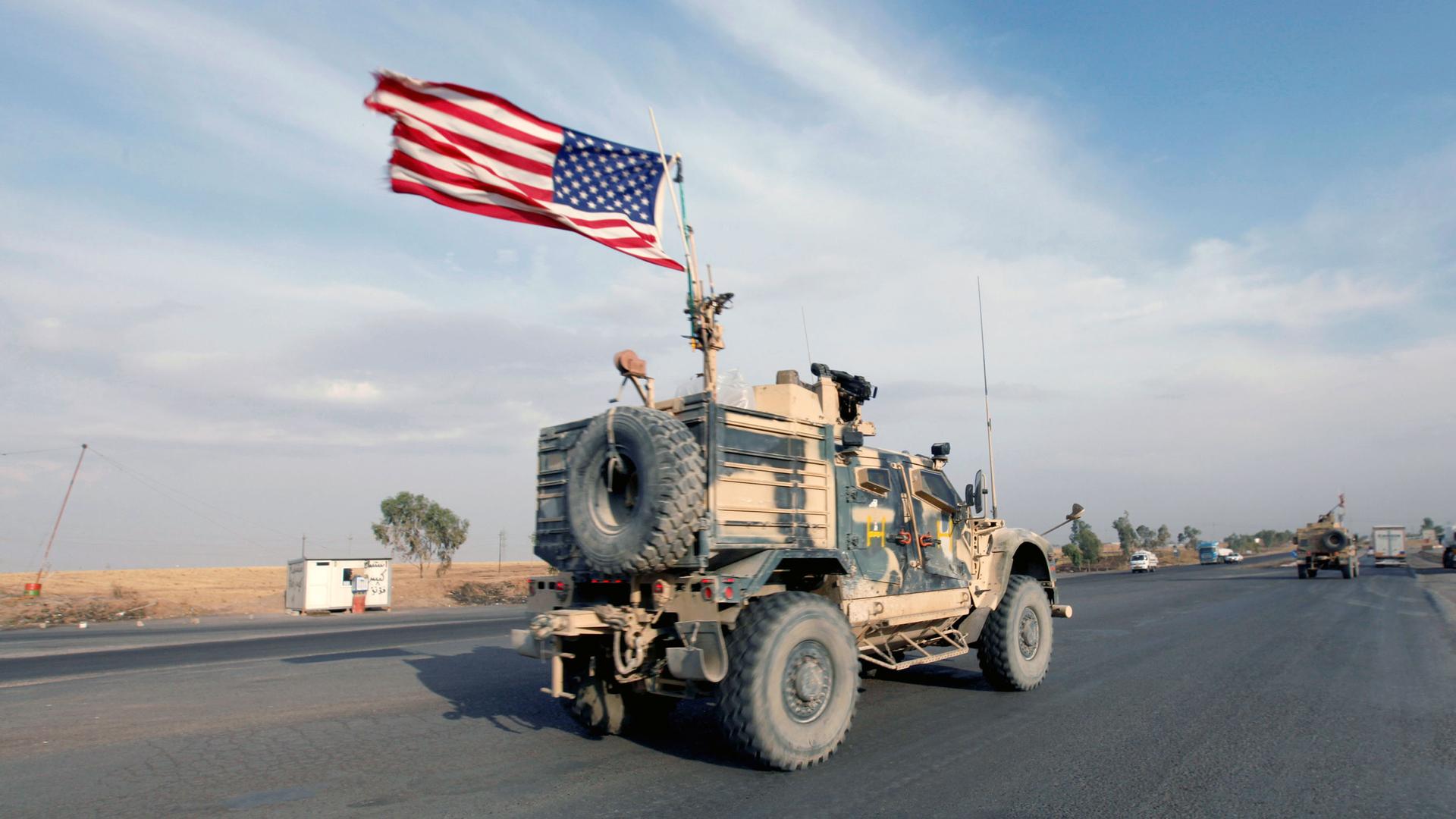 A military truck drives down a road with an American flag blowing in the wind on top of the truck